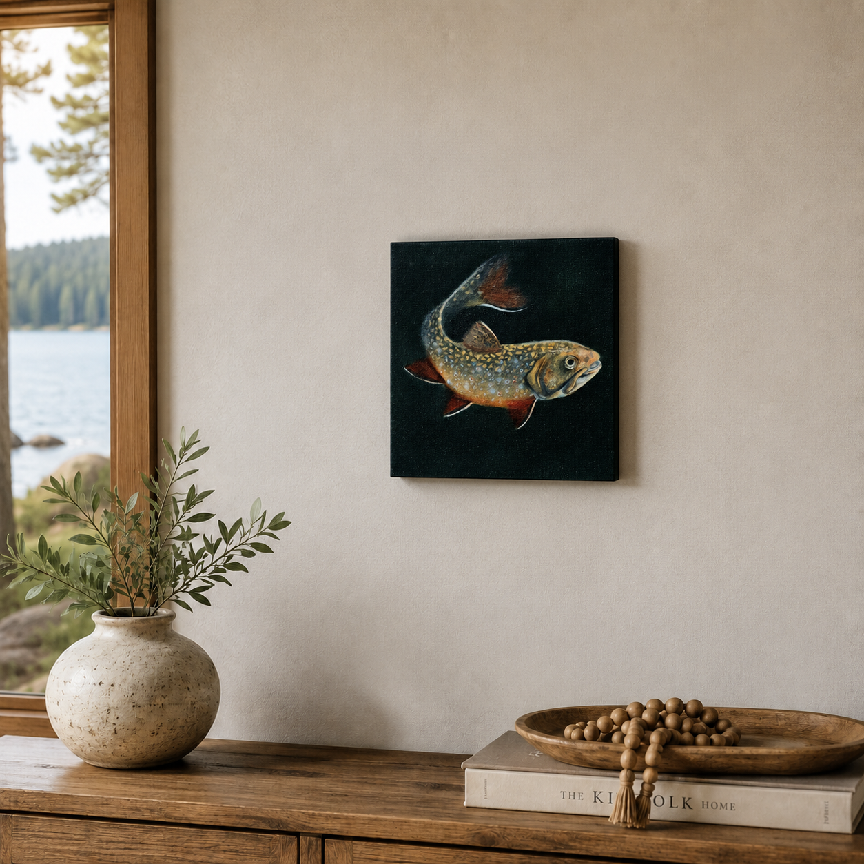 Small brook trout oil painting displayed in a luxury lake house setting above a wooden console with natural decor, books, greenery, and a lake view.
