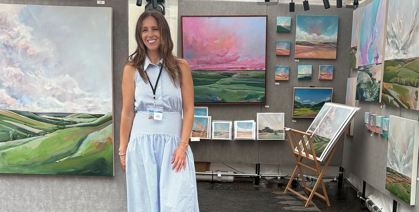 Woman smiling at an art exhibition, standing in front of landscape paintings on gray walls.