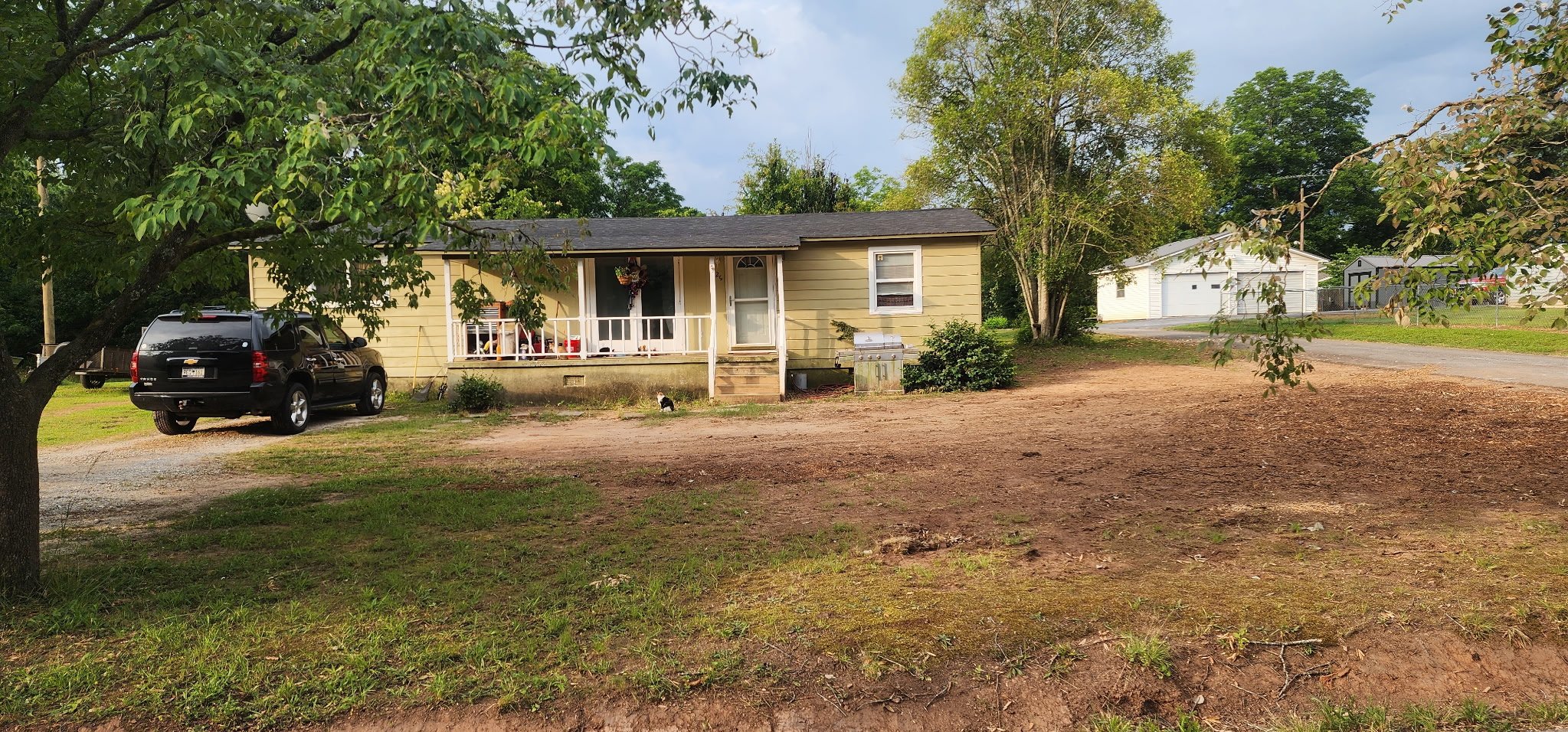 A small, yellow house with a front porch, surrounded by trees. A black SUV is parked in the driveway, and there's a small black and white cat near the steps. The yard is mostly dirt and grass.