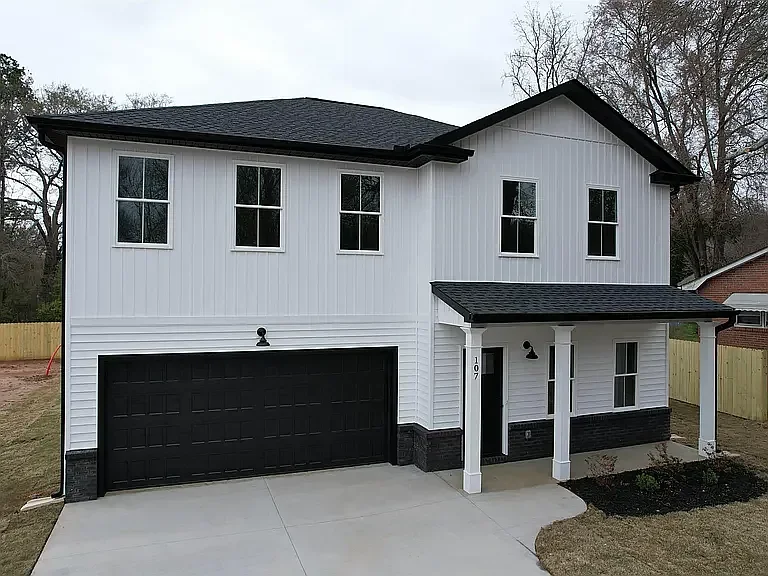 New two-story white house with black garage door and black roof, front porch with white columns, and a small landscaped area in front, surrounded by a wooden fence.
