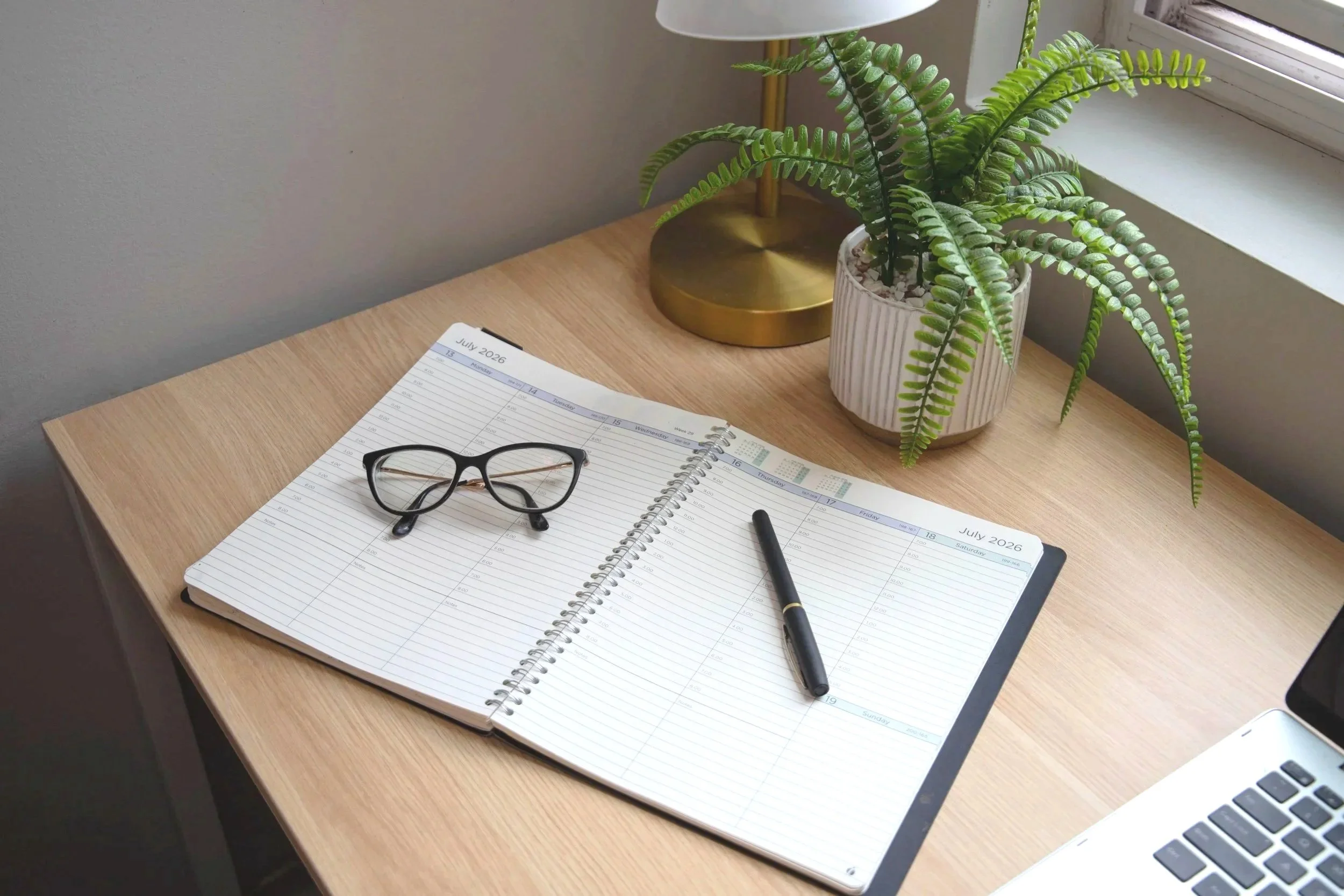 A wooden desk with a pair of black glasses, an open spiral planner with blank lined pages marked July 2026, a black pen, a white and gold table lamp, and a potted fern plant near a window.
