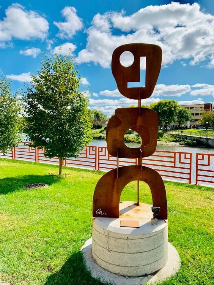 An abstract outdoor sculpture with rust-colored geometric shapes stacked vertically on a concrete base, with a tree and a river in the background under a partly cloudy sky.
