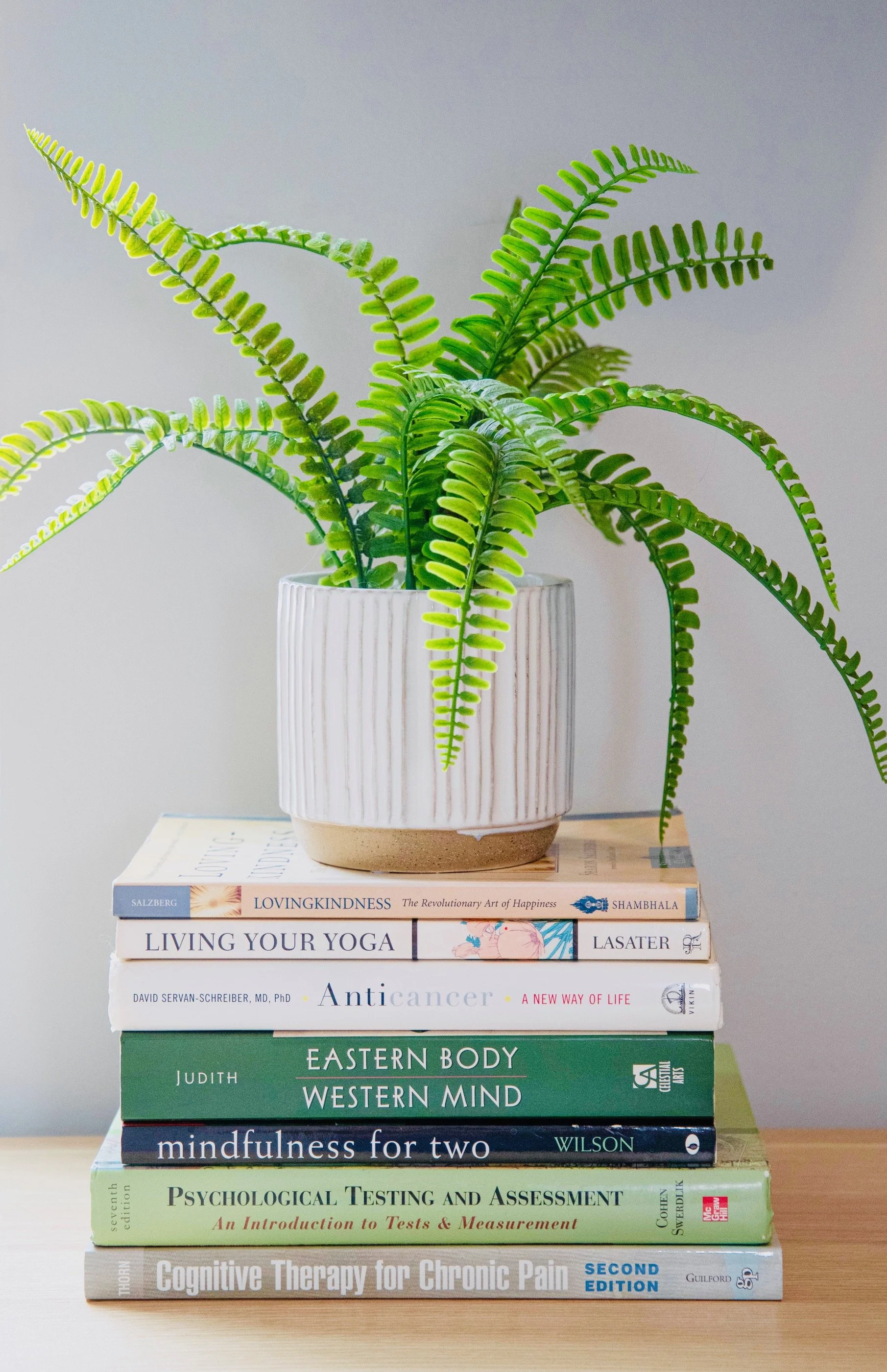 Stack of medical, wellness, and psychology books topped with a green fern plant in a white ribbed ceramic pot.