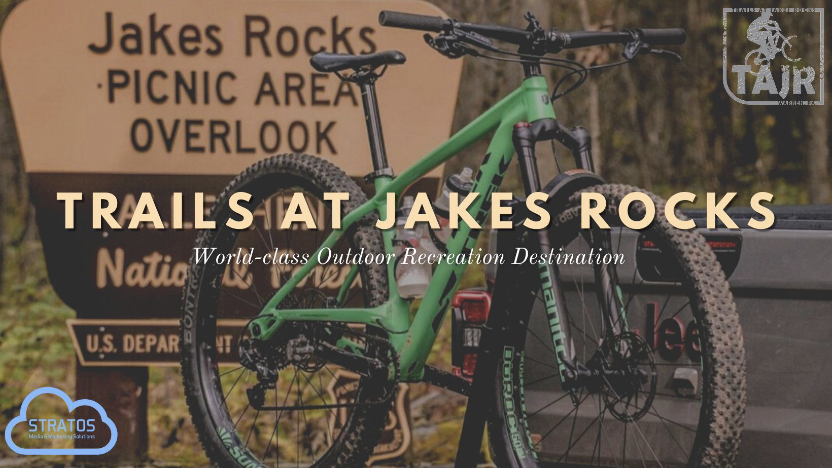 A green mountain bike leaning against a gray trailer with a Jakes Rocks sign in the background, promoting trails at Jakes Rocks as a world-class outdoor recreation destination.