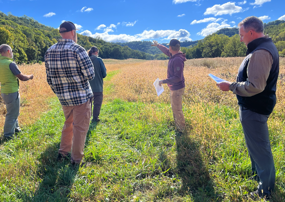 Six people standing in a field during daytime, engaged in a discussion about trail planning, surrounded by green trees and hills under a partly cloudy sky.