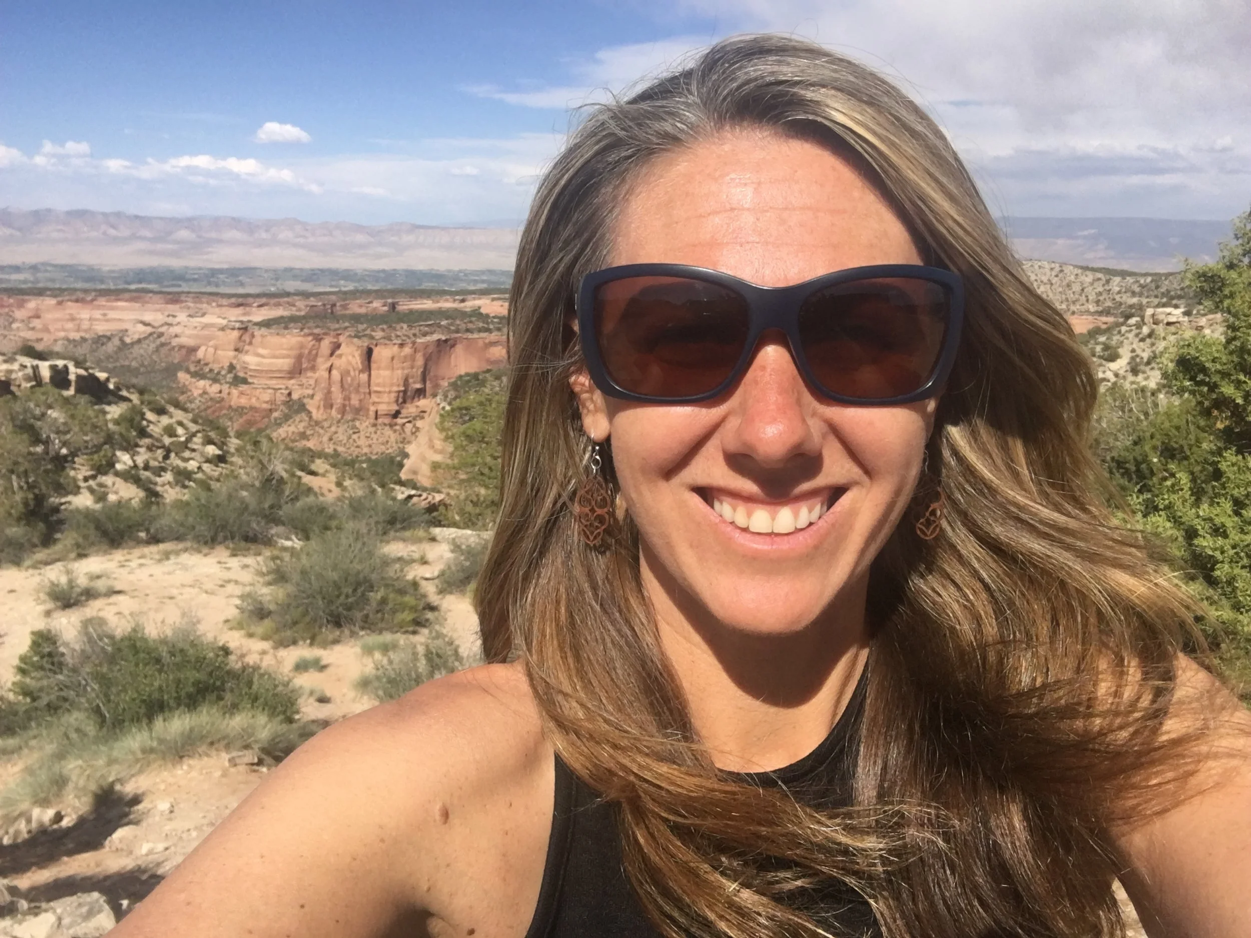 Aaryn Kay, taking a selfie outdoors with a canyon and desert landscape in the background under a partly cloudy sky.