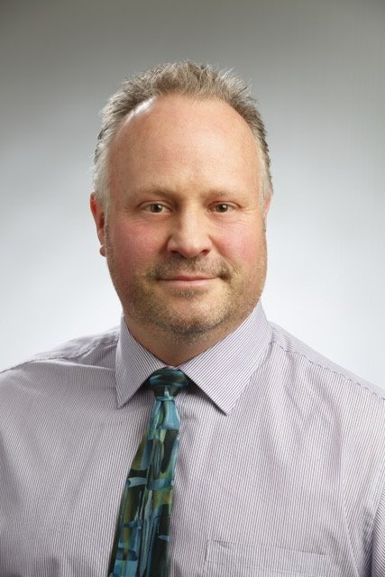 A man with short, curly, light-colored hair, wearing a light purple striped dress shirt and a blue patterned tie, posing against a plain gray background.
