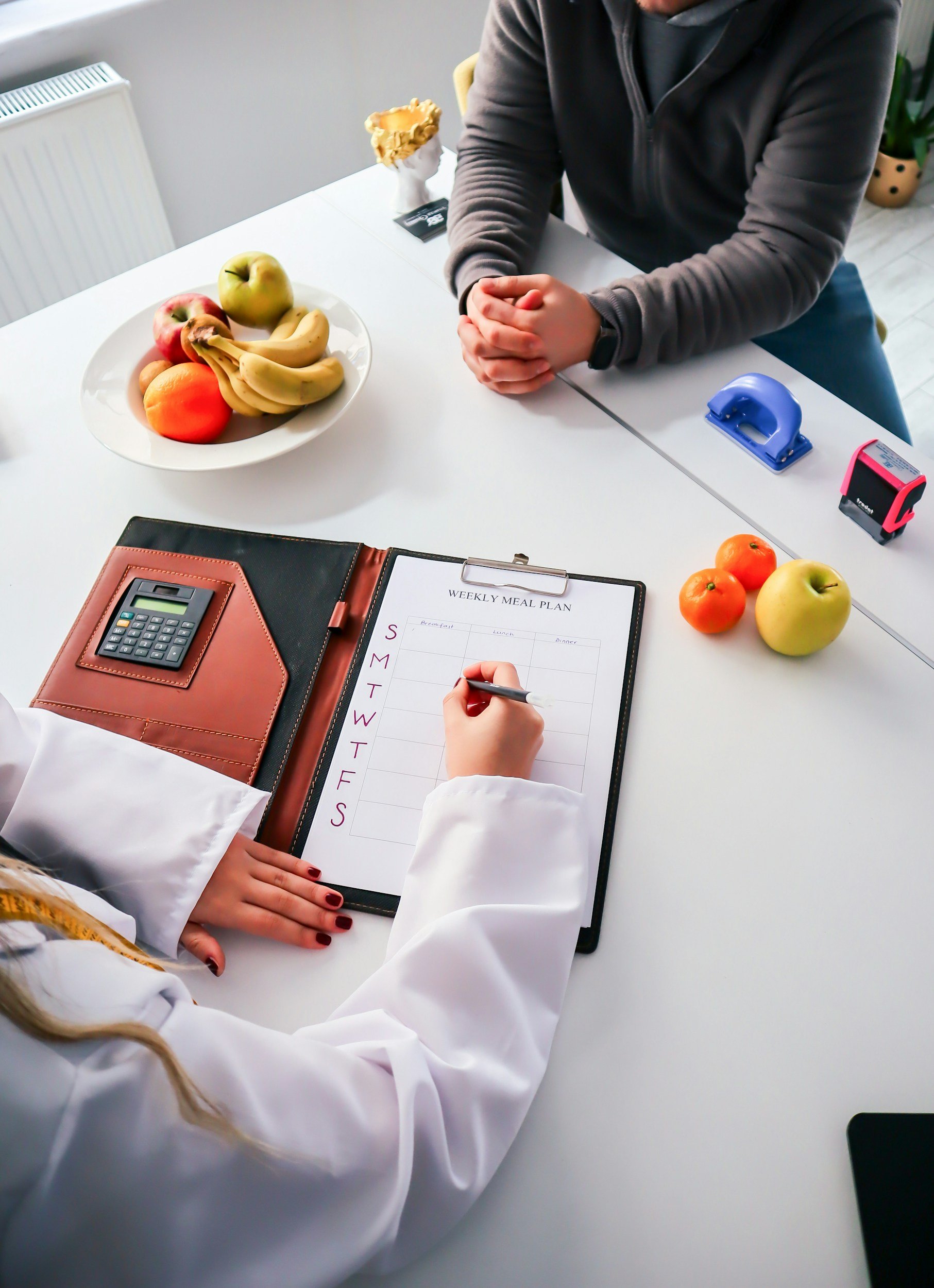 Person with long blonde hair in a white shirt sitting at a white table, writing in a weekly meal plan notebook with a black pen. On the table, there is a brown leather folder with a calculator attached, a plate with bananas, apples, and an orange, two small tomatoes, a blue hole puncher, a pink and black tape dispenser, and a black smartphone. In the background, there is a small potted plant and a figurine with a golden crown.