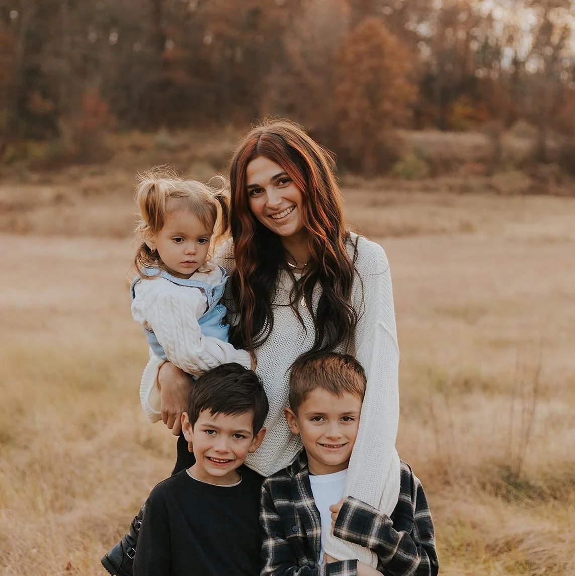 A woman with long dark hair smiling, standing in a field with three children. Two boys are in front, one girl perched on her arm, all smiling or looking serious. Autumn trees in the background.