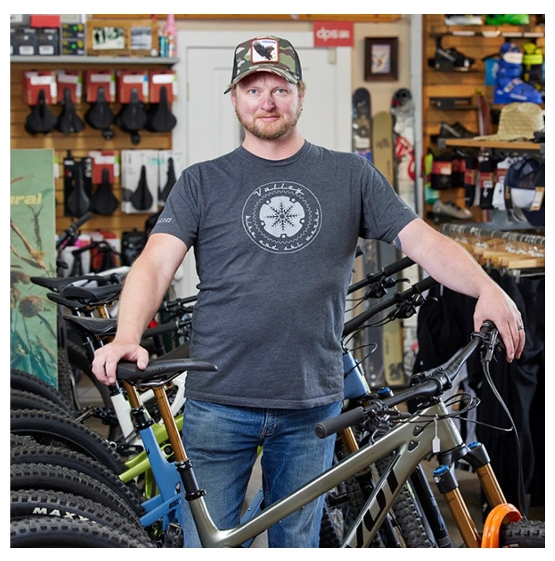 A man standing inside a bike shop, holding a mountain bike, surrounded by other bikes and cycling accessories.