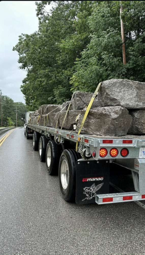 A flatbed truck carrying large rocks on a wet road, with green trees and utility poles in the background.