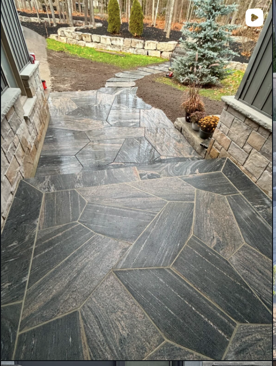 A stone patio with a geometric pattern, leading to a garden with a pathway, small trees, and potted plants on the side.