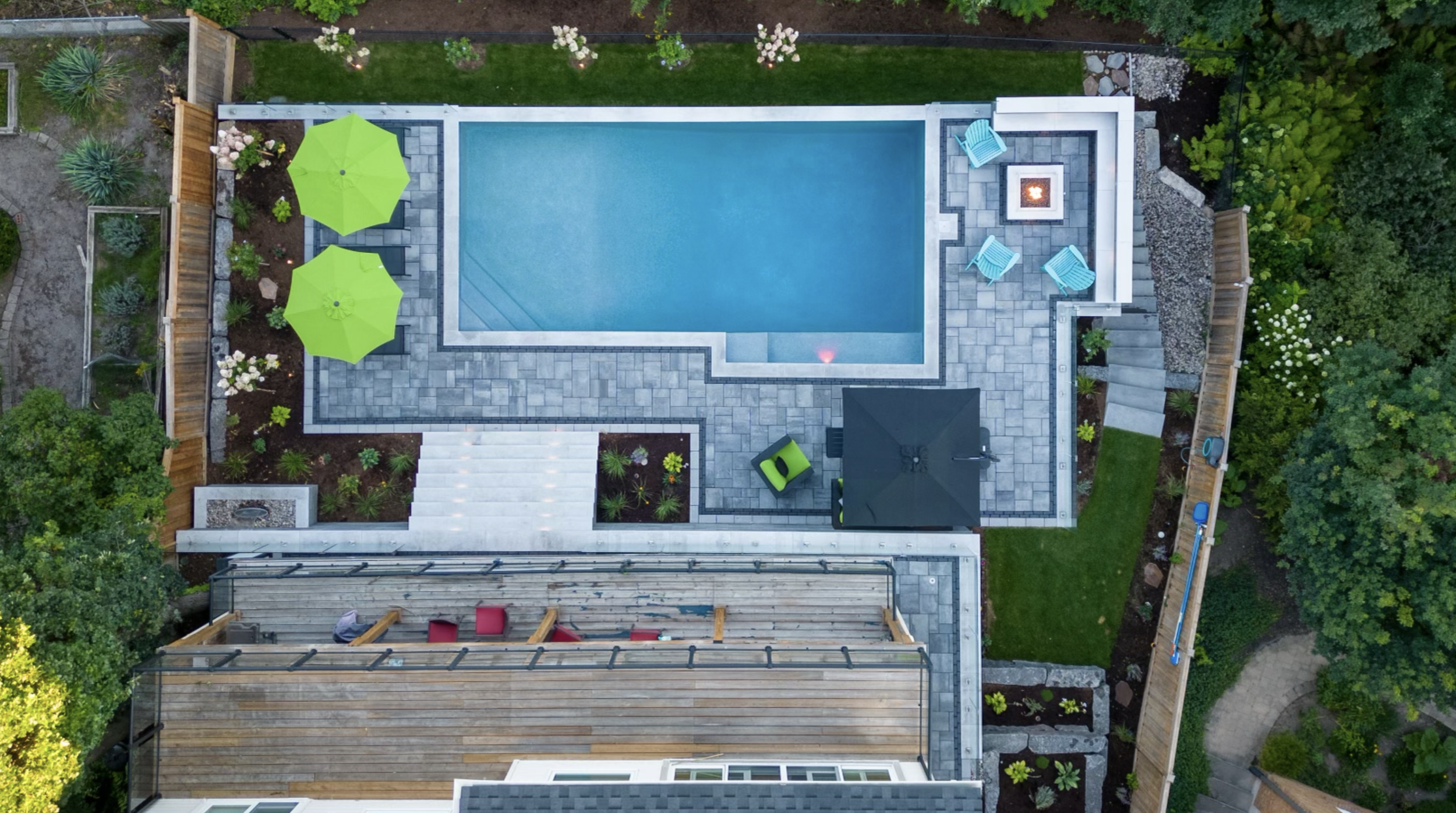 A backyard with a swimming pool, surrounded by a stone patio, green grass, and landscaped garden beds. There are two lime green umbrellas, a fire pit with chairs, and a seating area with red cushions. Trees and plants border the yard.