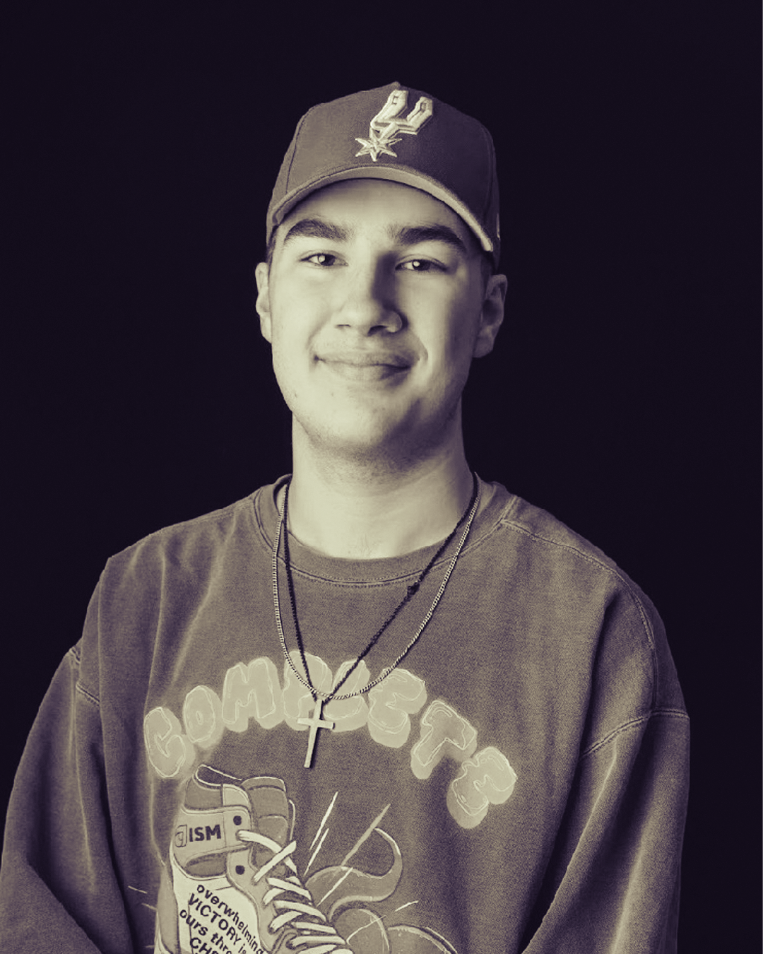 Black and white photo of a young man wearing a baseball cap, multiple necklaces, and a graphic t-shirt, smiling against a dark background.