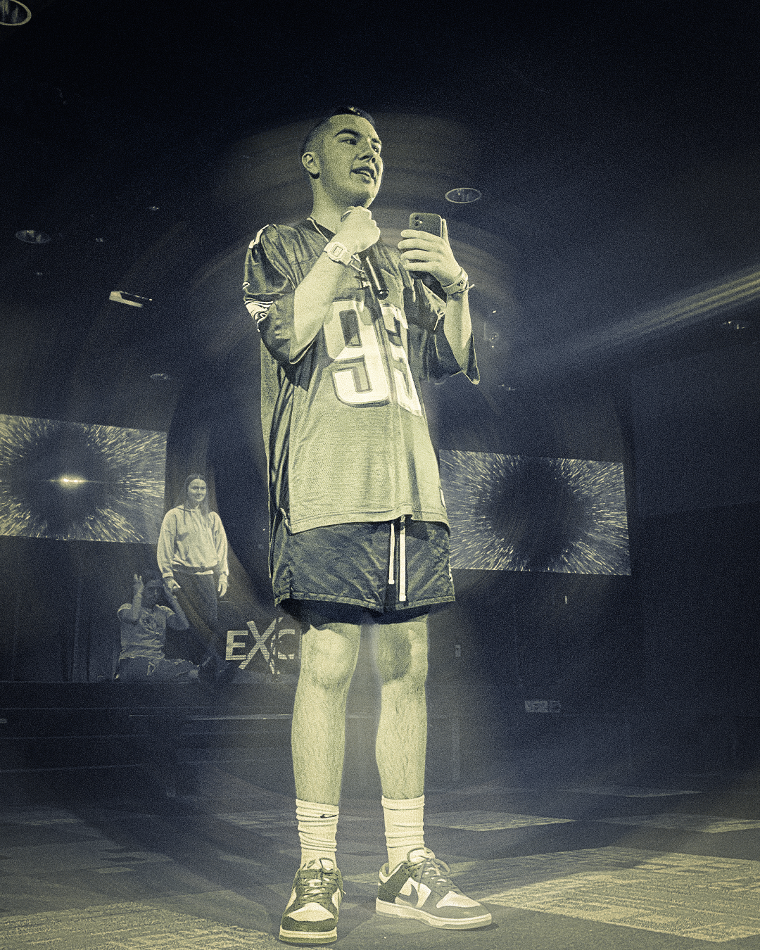 A young man in sportswear taking a selfie with a smartphone on stage, with two large screens and a woman in the background.