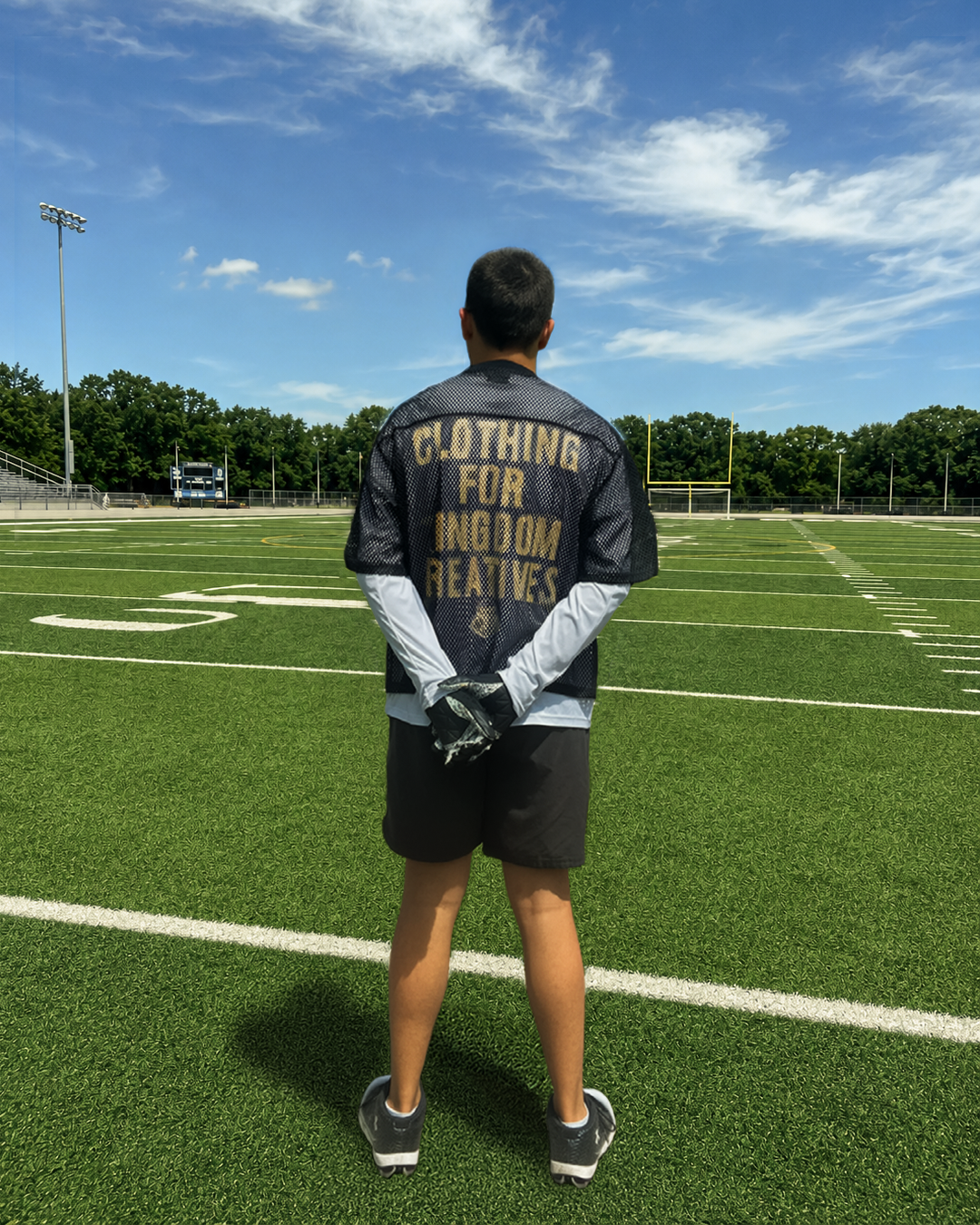 Young male football player standing on a football field, facing away, wearing a black mesh jersey with gold lettering that says 'Clothing for ingrown reactive,' holding gloves behind his back, with a clear blue sky overhead and a goal post in the background.