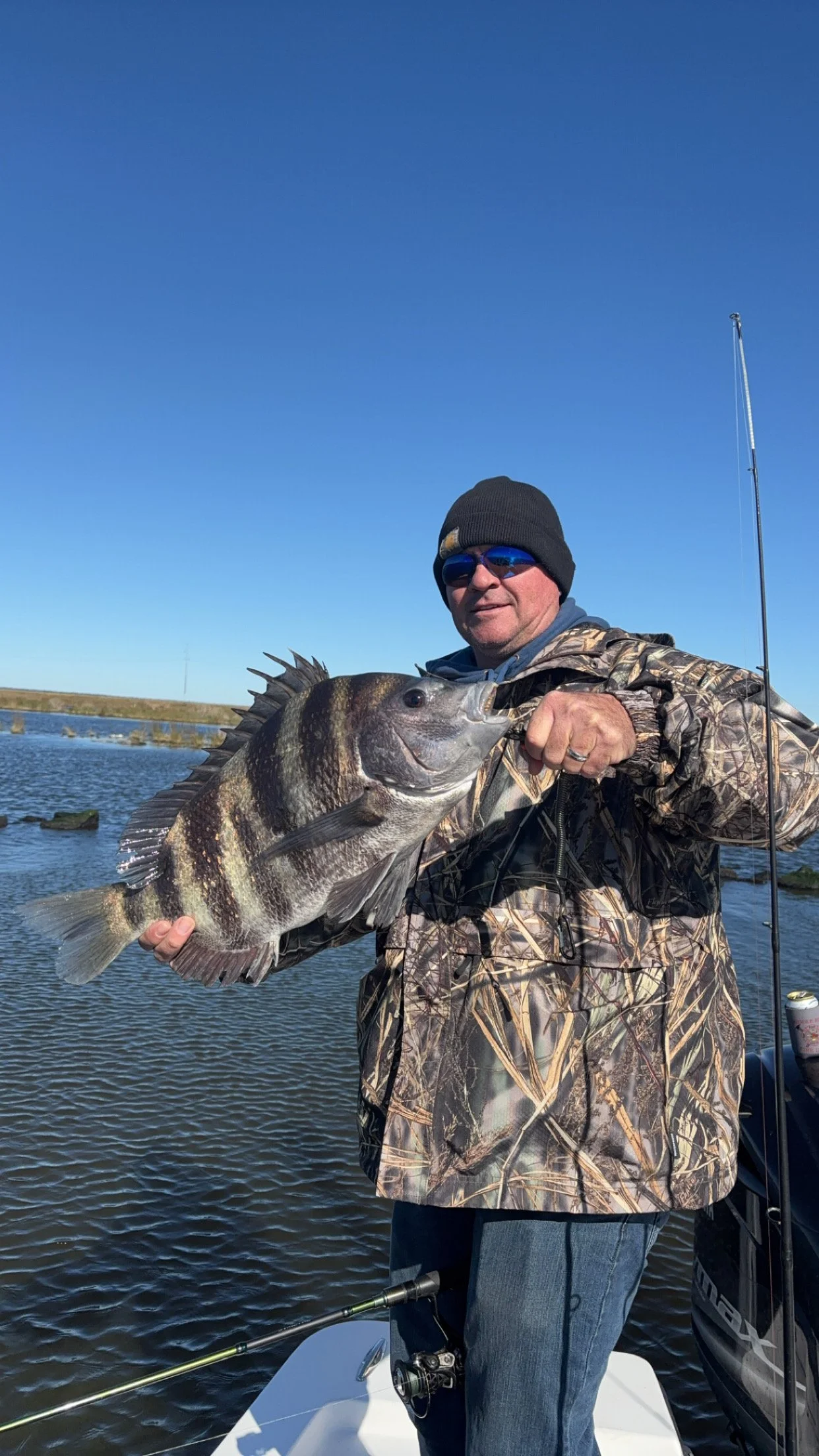 A man wearing camouflage jacket, black beanie, and sunglasses holding a large fish on a boat in a lake with blue sky in the background.