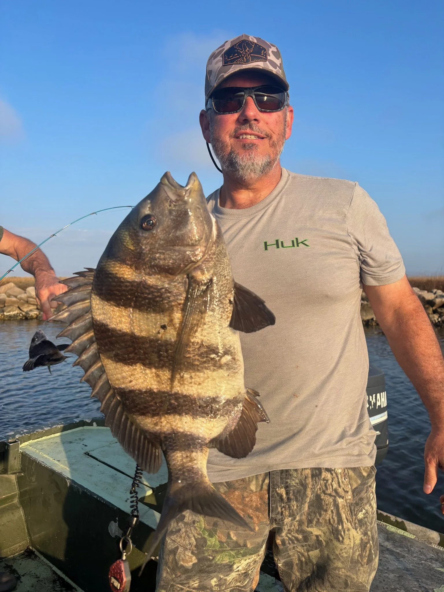 A man holding a large fish on a boat near a rocky shoreline, wearing sunglasses, a baseball cap, a gray t-shirt with 'Huk' written on it, and camo pants, with a blue sky background.