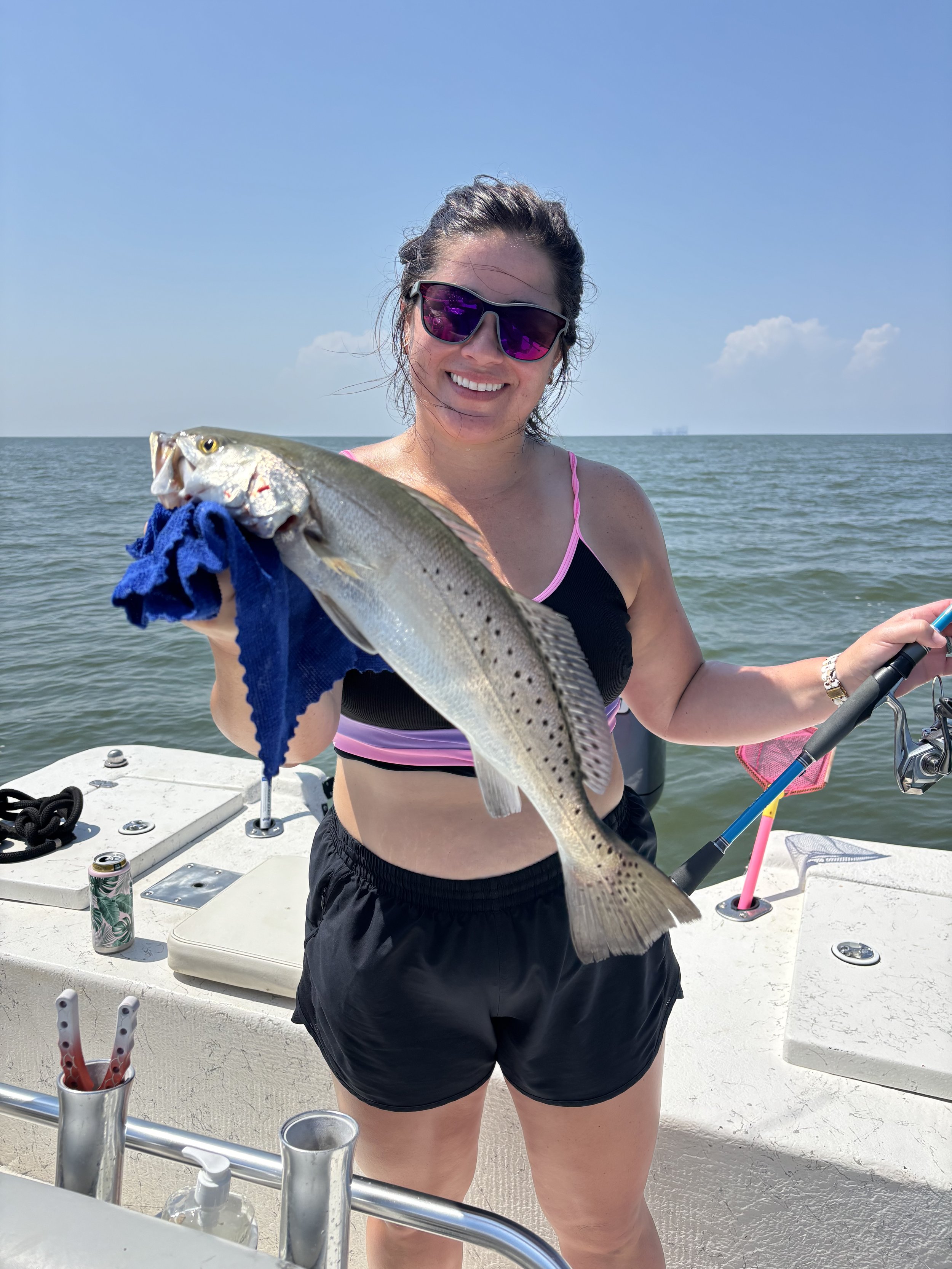 Woman with dark hair, wearing sunglasses and a black and pink tank top, holding a large fish on a boat at the water.