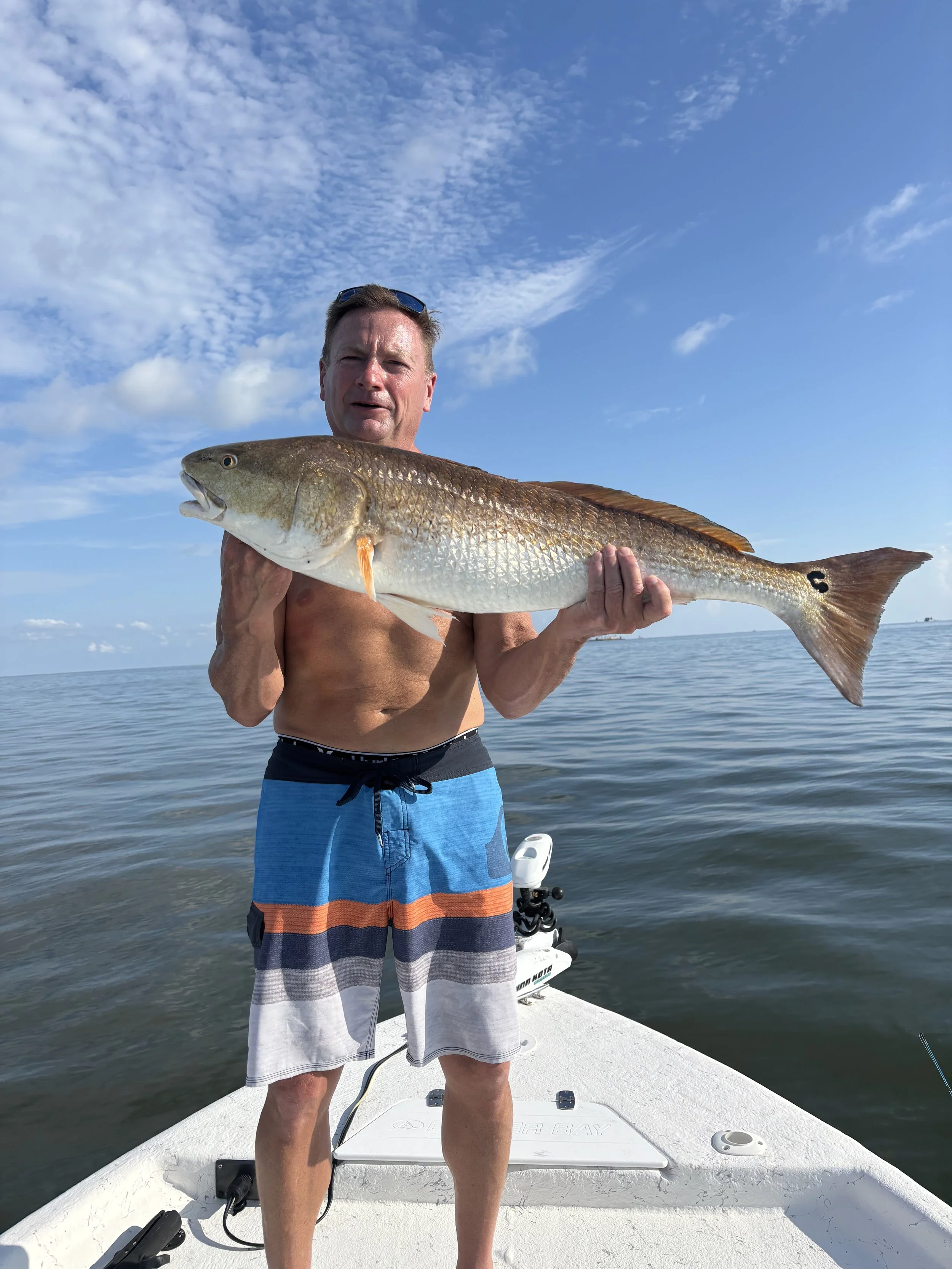 A man in swim shorts holding a large fish on a boat in the water. The sky is partly cloudy and the man appears to be on a fishing trip.