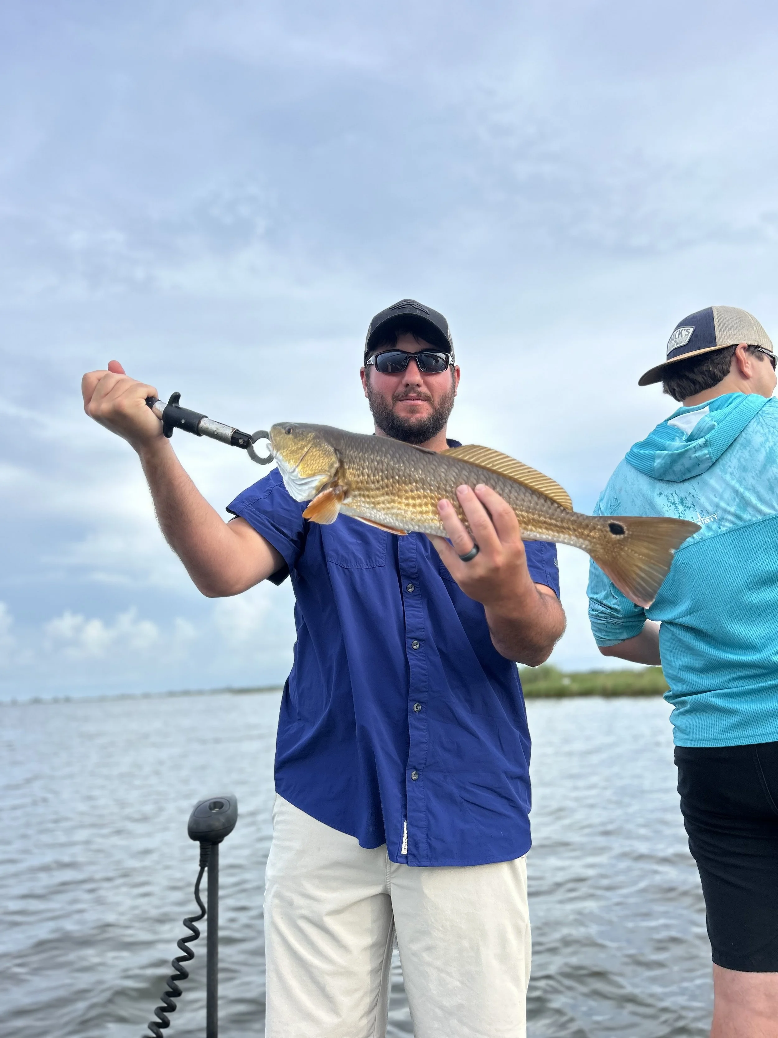 A man holding a fish on a boat, with another person partially visible in the background, water and sky in the background.