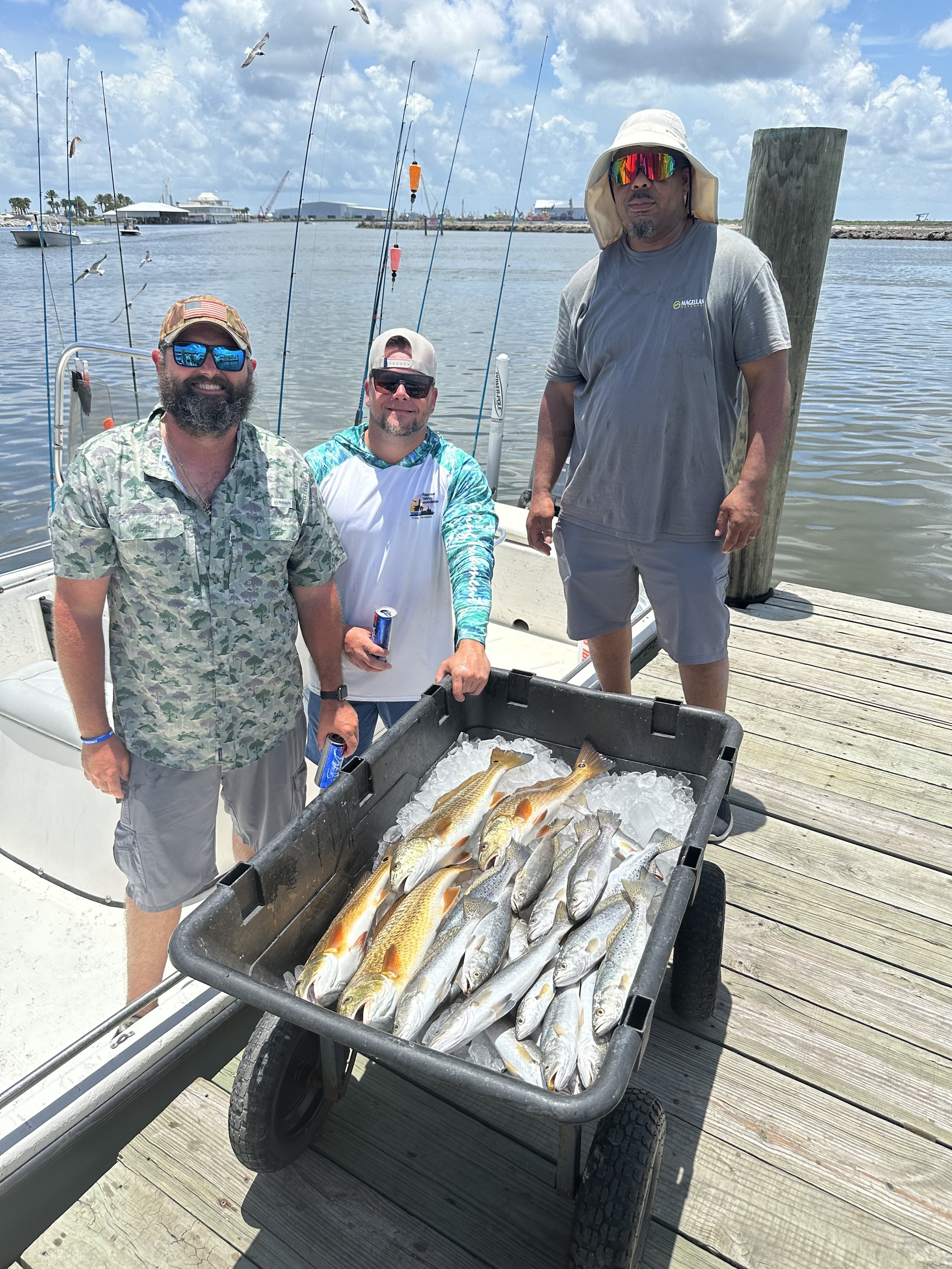 Three men standing on a dock with a boat in the background. In front of them is a cart filled with caught fish. The men are smiling, wearing casual fishing attire and sunglasses, with a partly cloudy sky and water in the background.