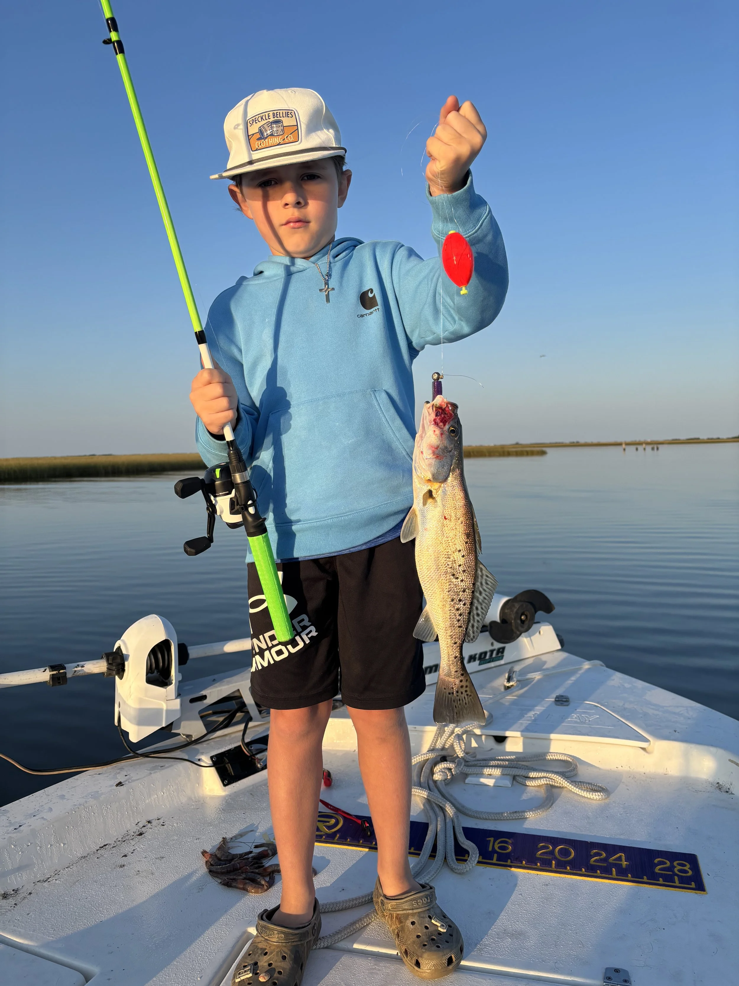 A boy standing on a boat holding a large fish he caught while fishing. The boy is wearing a light blue hoodie, a beige baseball cap, and Crocs. The boat is on a calm body of water with a clear blue sky, and fishing gear, including a rod, and a measur