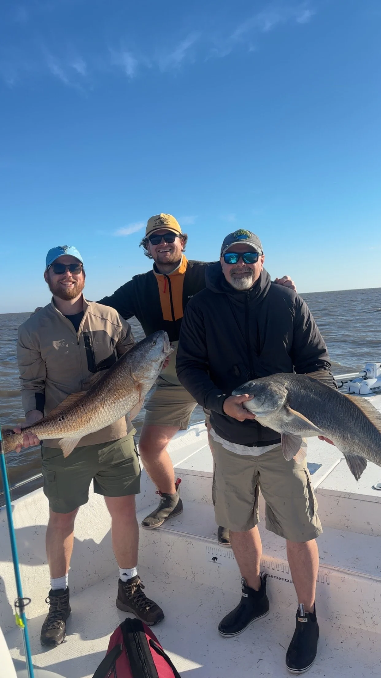 Three men on a boat holding large fish they caught while fishing, with a body of water and blue sky in the background.