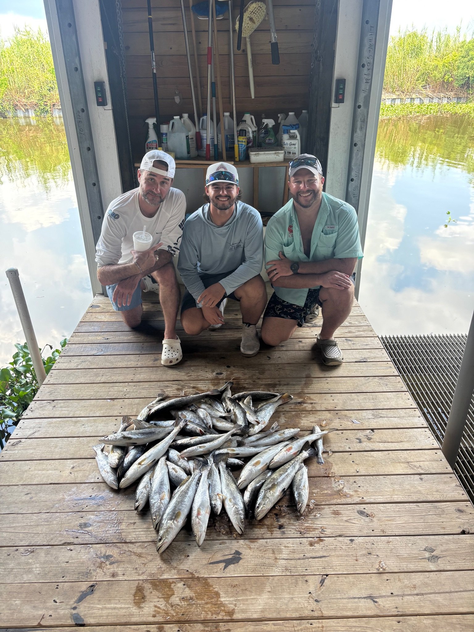 Three men squatting on a wooden dock in front of a pile of fish, with fishing gear and supplies on a rack behind them and water on either side.