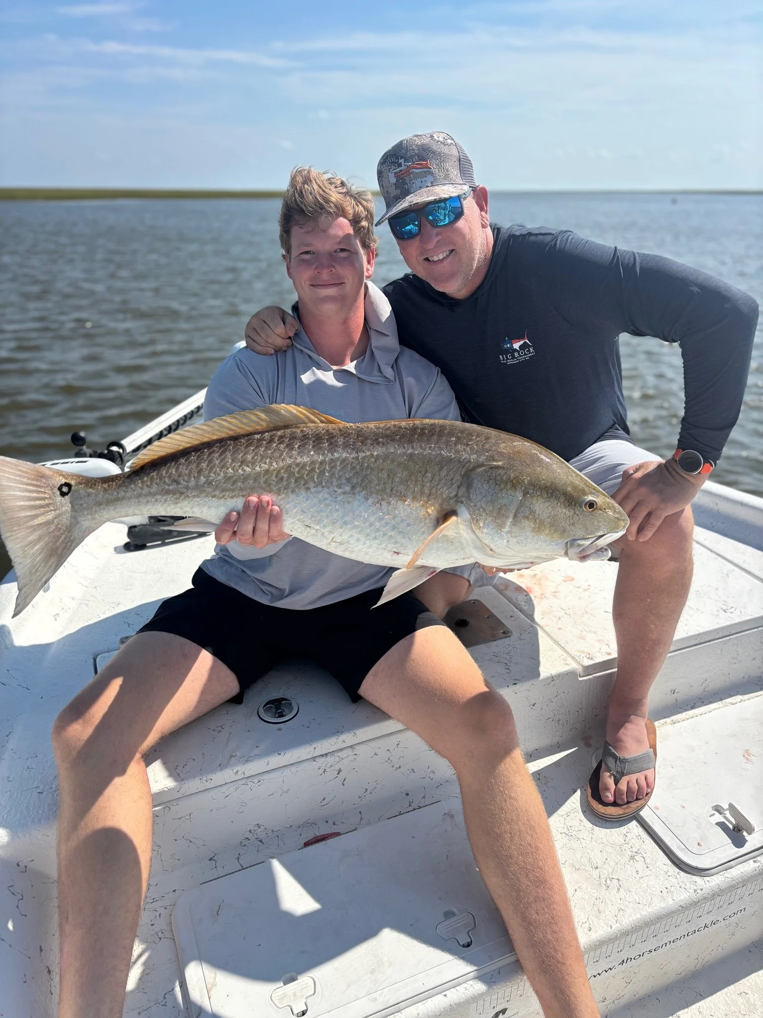 Two men on a boat holding a large fish, smiling, with water and a cloudy sky in the background.