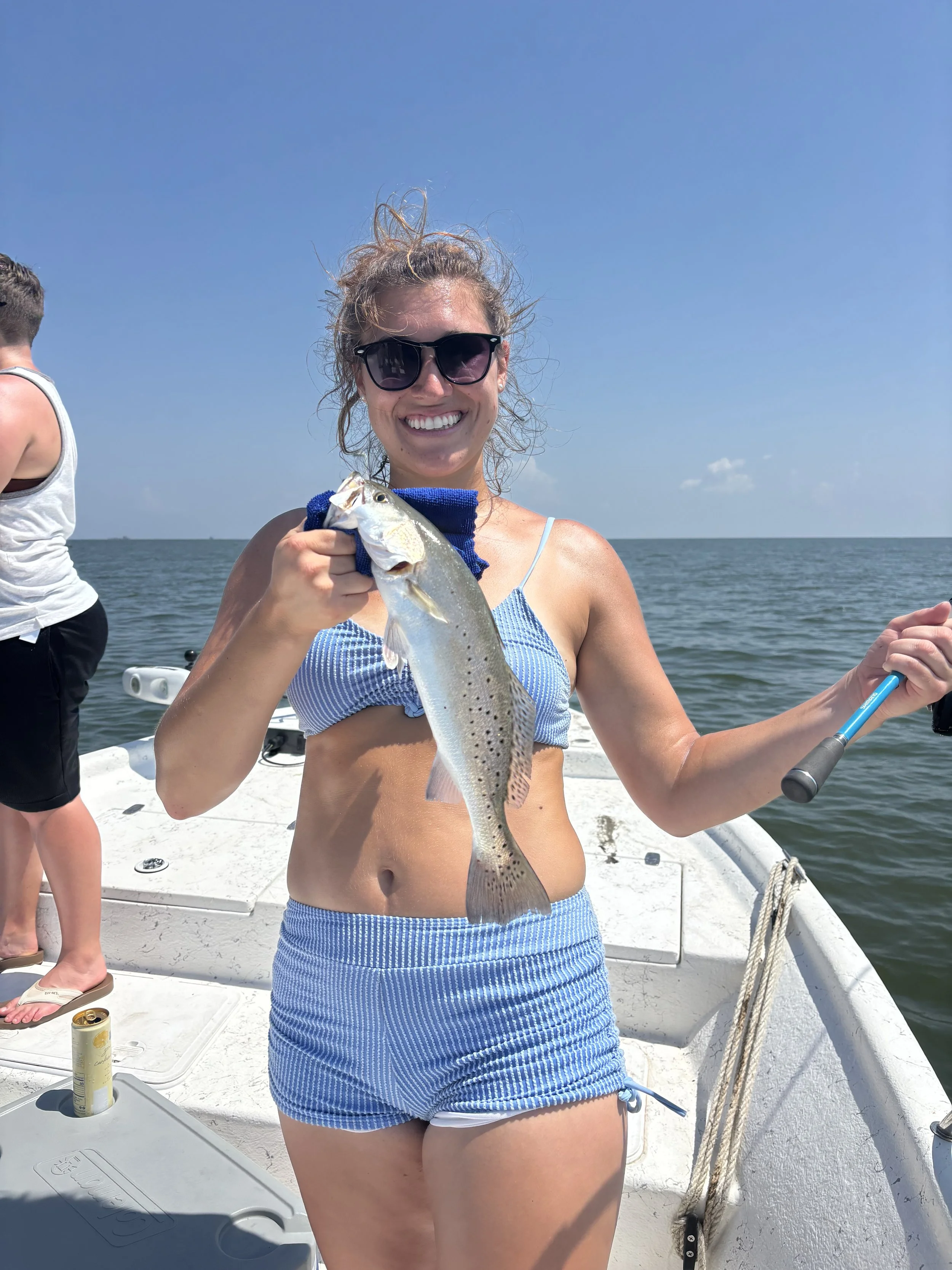 A smiling woman wearing sunglasses and a blue and white striped two-piece swimsuit holding a fish on a boat in the water on a sunny day.