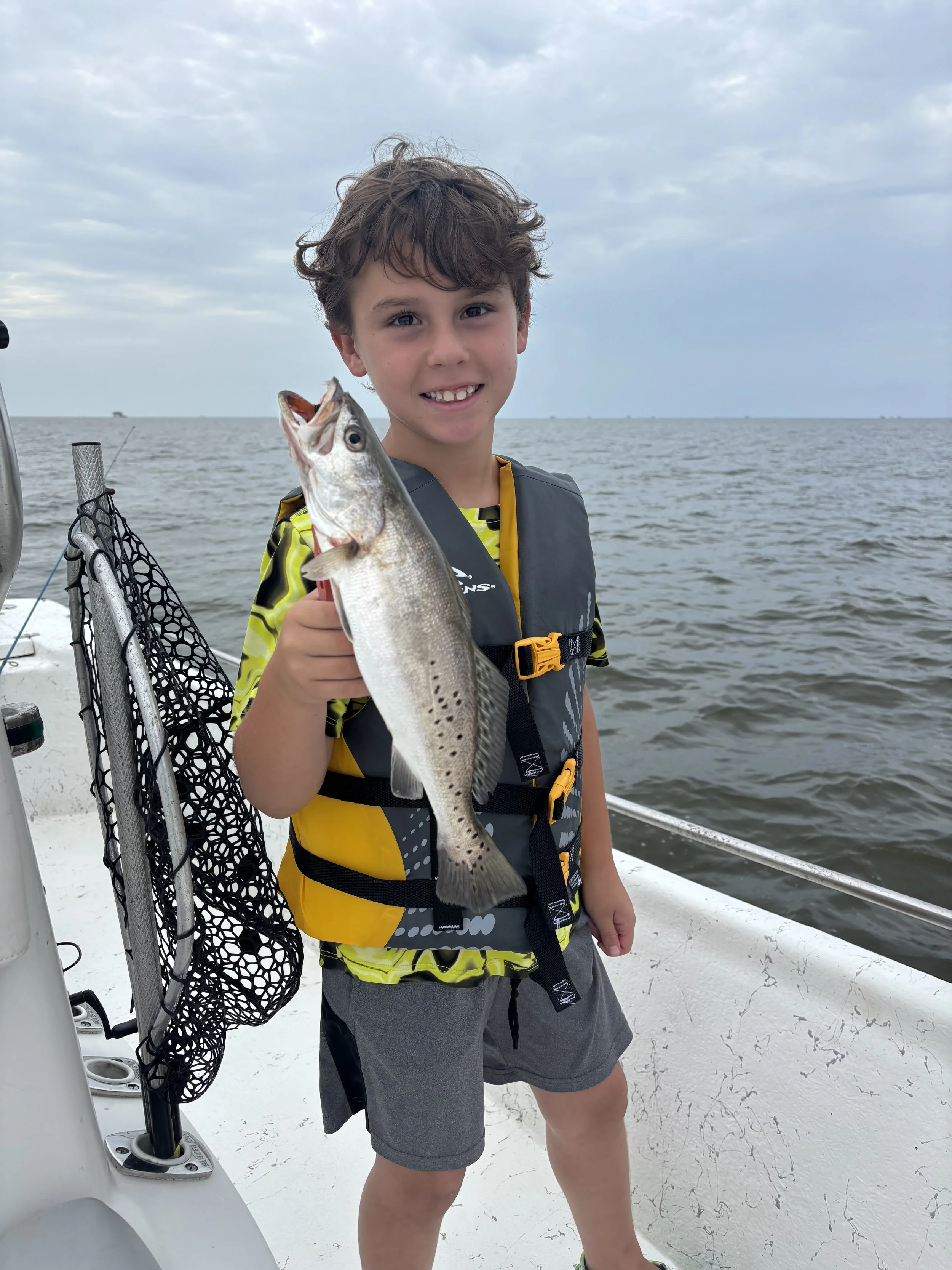 A young boy with curly hair, smiling, holding a large fish on a boat in a body of water with a cloudy sky in the background.
