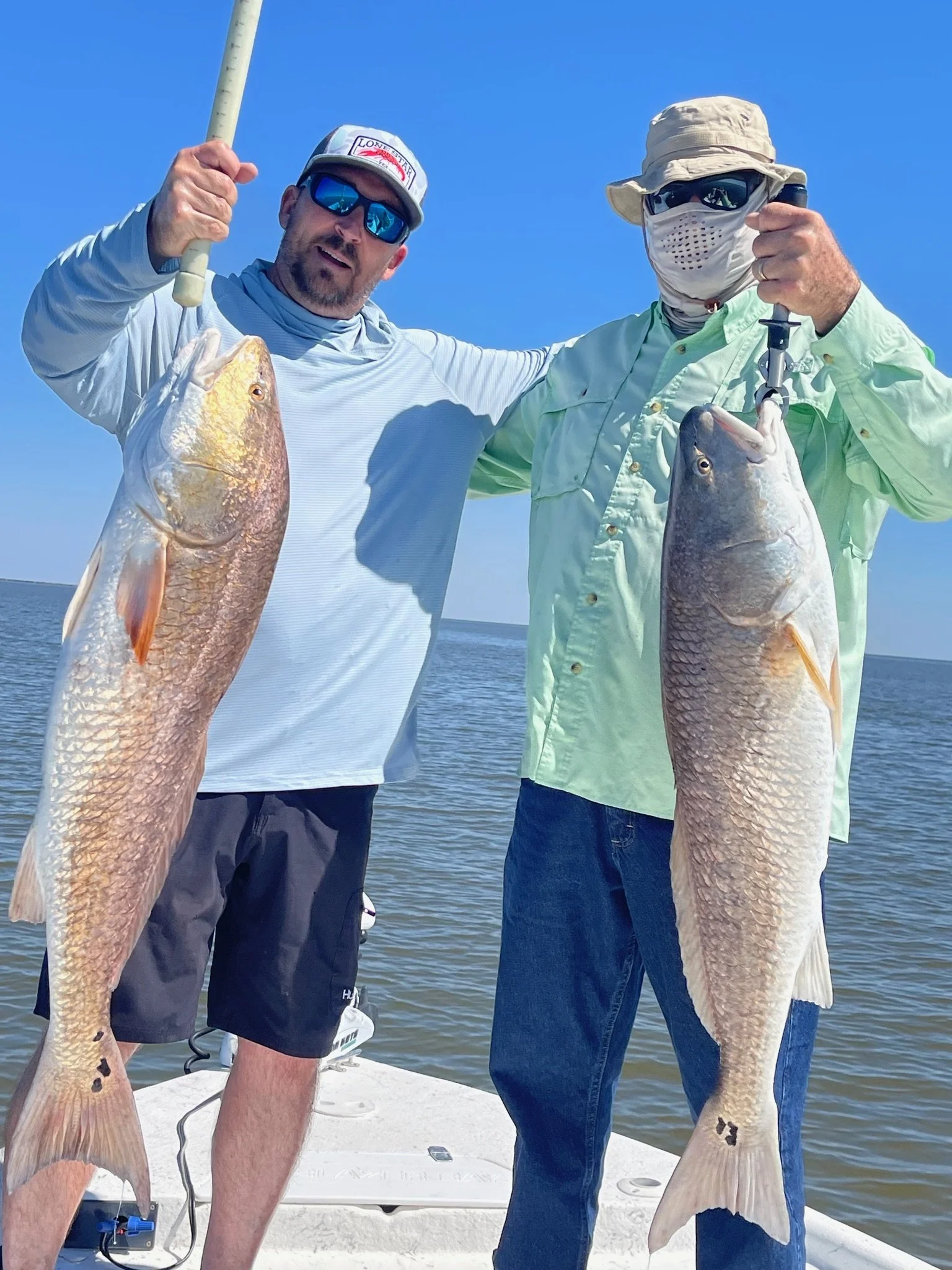 Two men holding large fish on a boat, with water and a clear blue sky in the background.