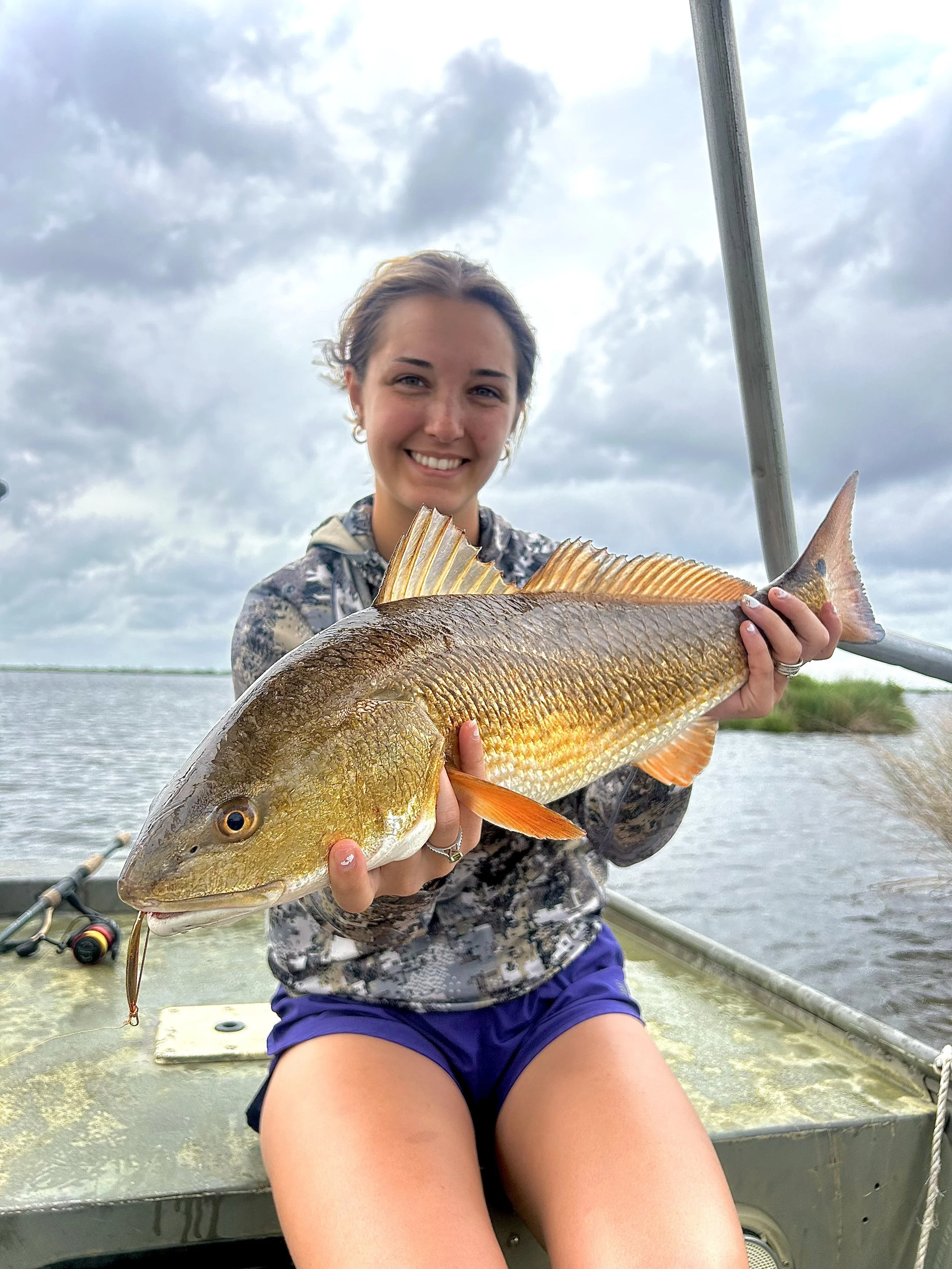 A woman smiling and holding a large fish on a boat in a body of water with cloudy sky in the background.