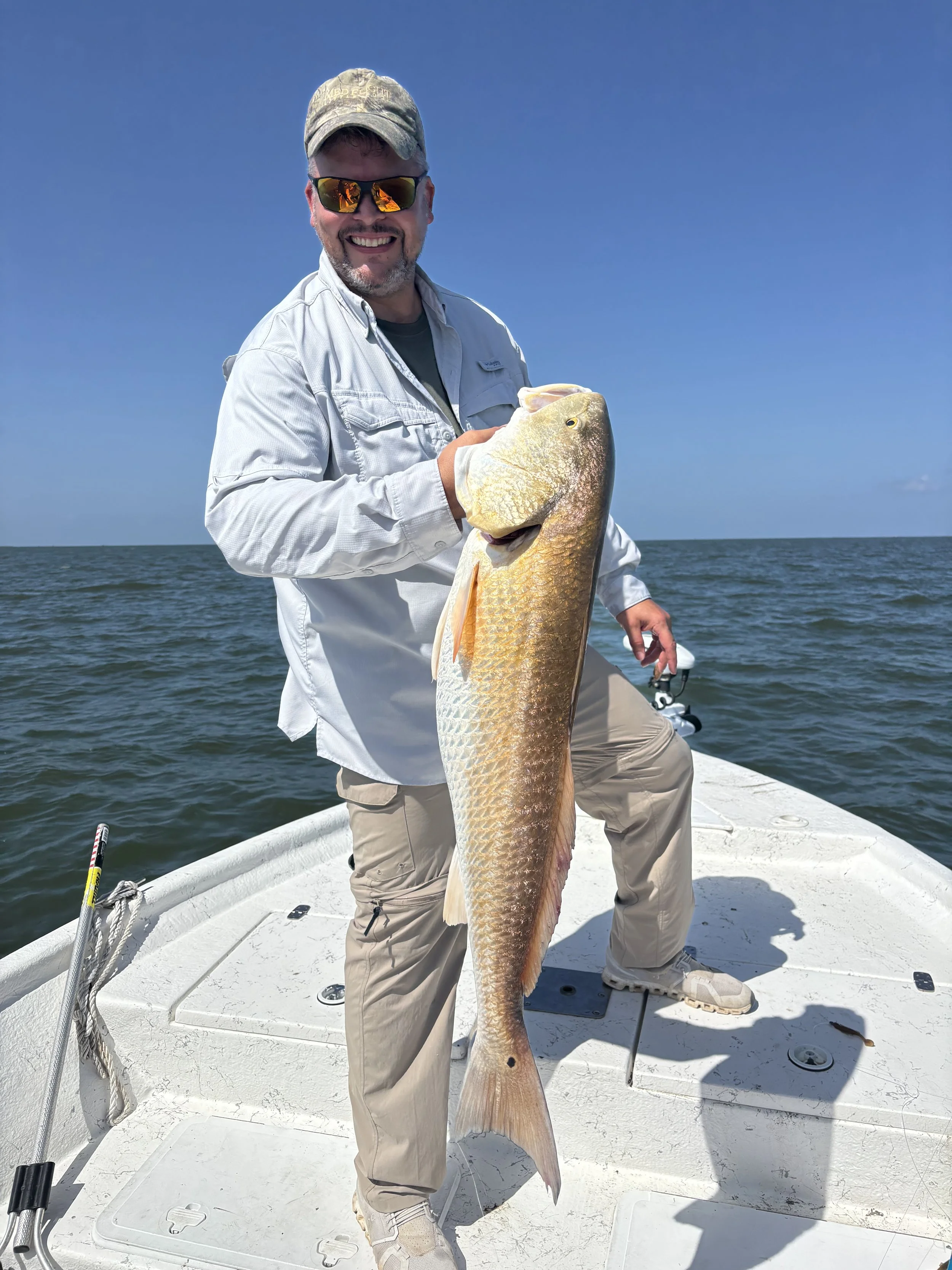 A man standing on a boat holding a large fish, with a wide smile, wearing sunglasses, a cap, and a long-sleeved shirt, on a body of water under a blue sky.