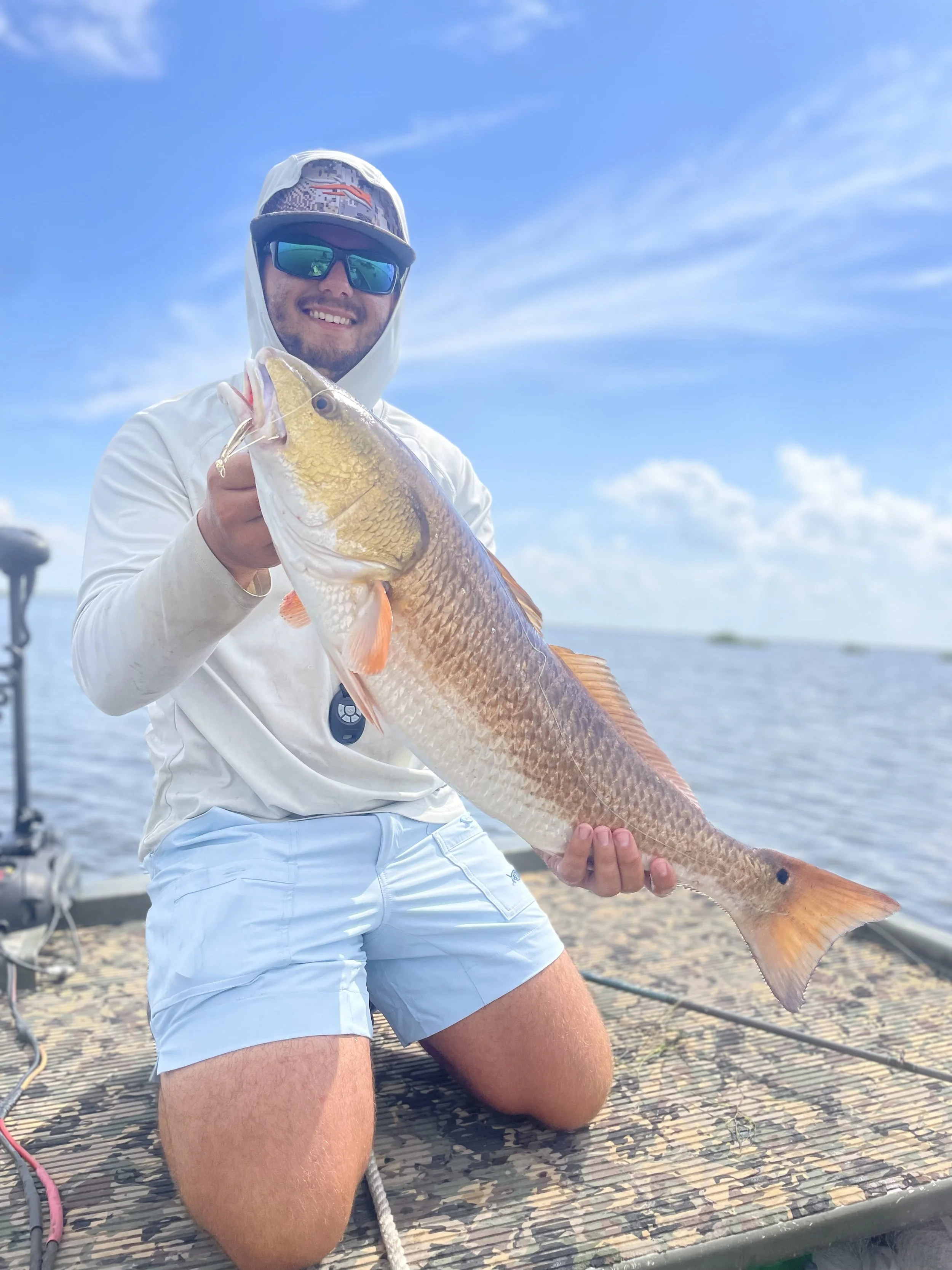A man kneeling on a fishing dock holding a large fish with a golden-yellow and grayish body. The man is smiling, wearing sunglasses, a white hoodie, and light blue shorts, and is outdoors under a partly cloudy sky.