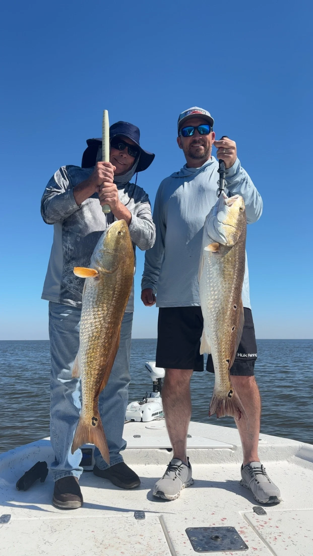Two men on a boat holding up large fish, smiling, with a clear blue sky and water in the background.