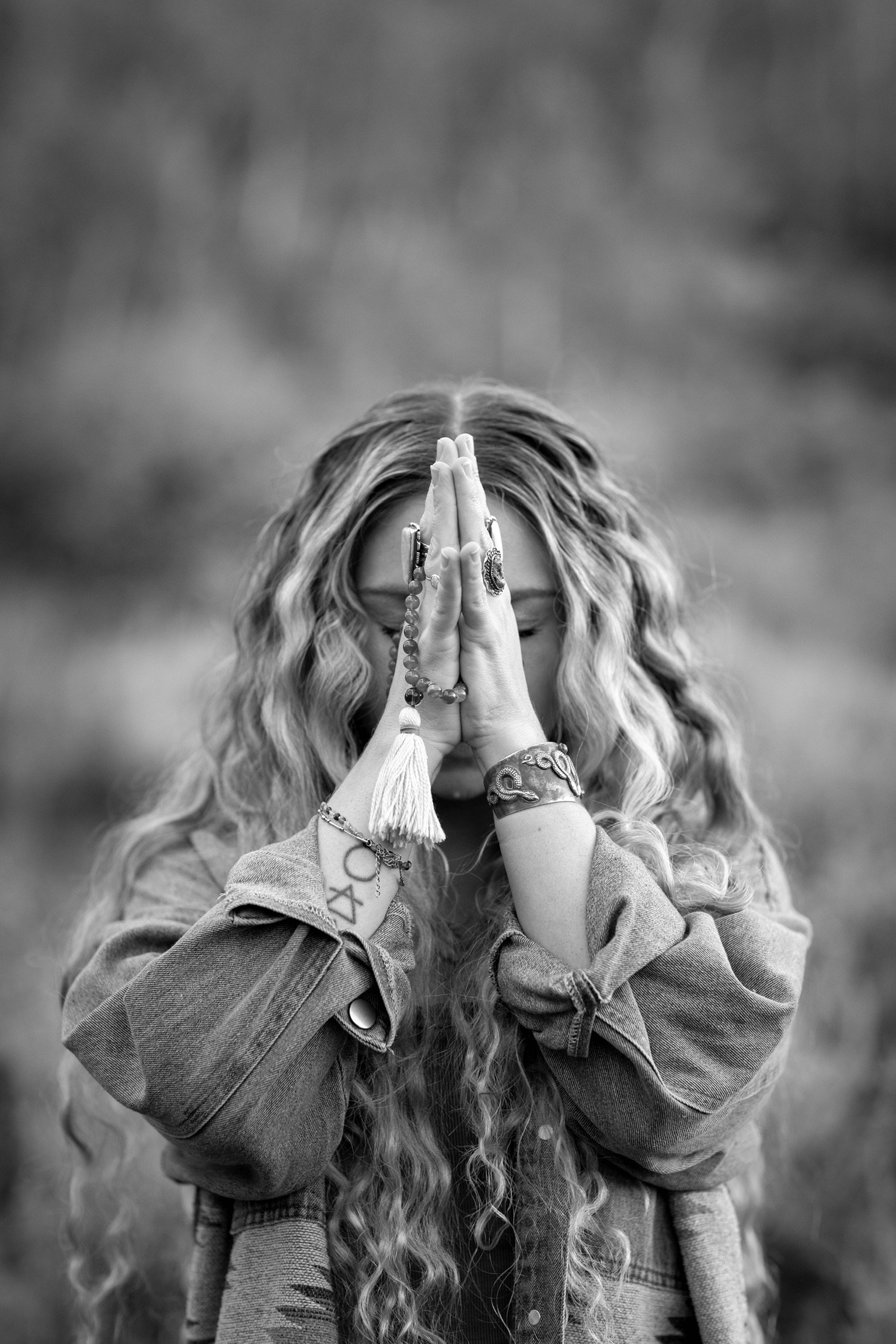 A woman with curly hair standing outdoors with her hands pressed together in a prayer position, wearing multiple rings, jewelry, and a denim jacket, in black and white.