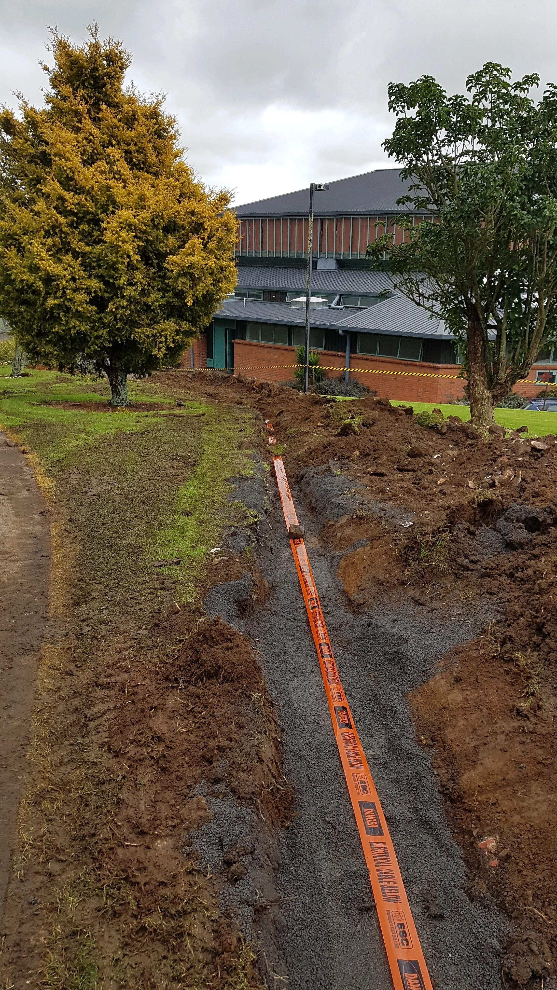 A trench dug into the ground with gravel inside, marked with an orange electrician safety tape placed by DB Electrical, in front of a modern building with trees and cloudy sky.