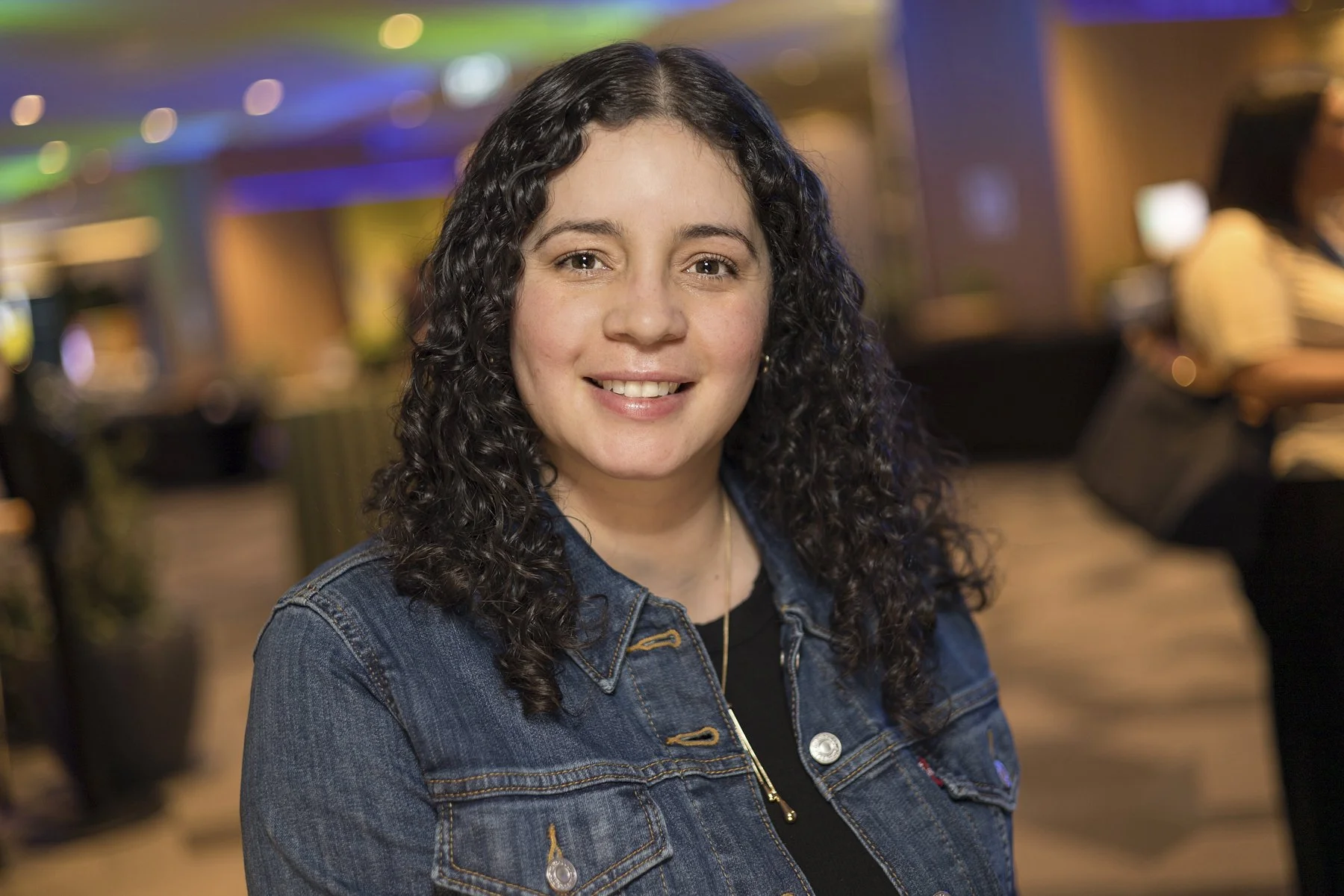 A woman with curly dark hair smiling at the camera in a warmly lit indoor setting.