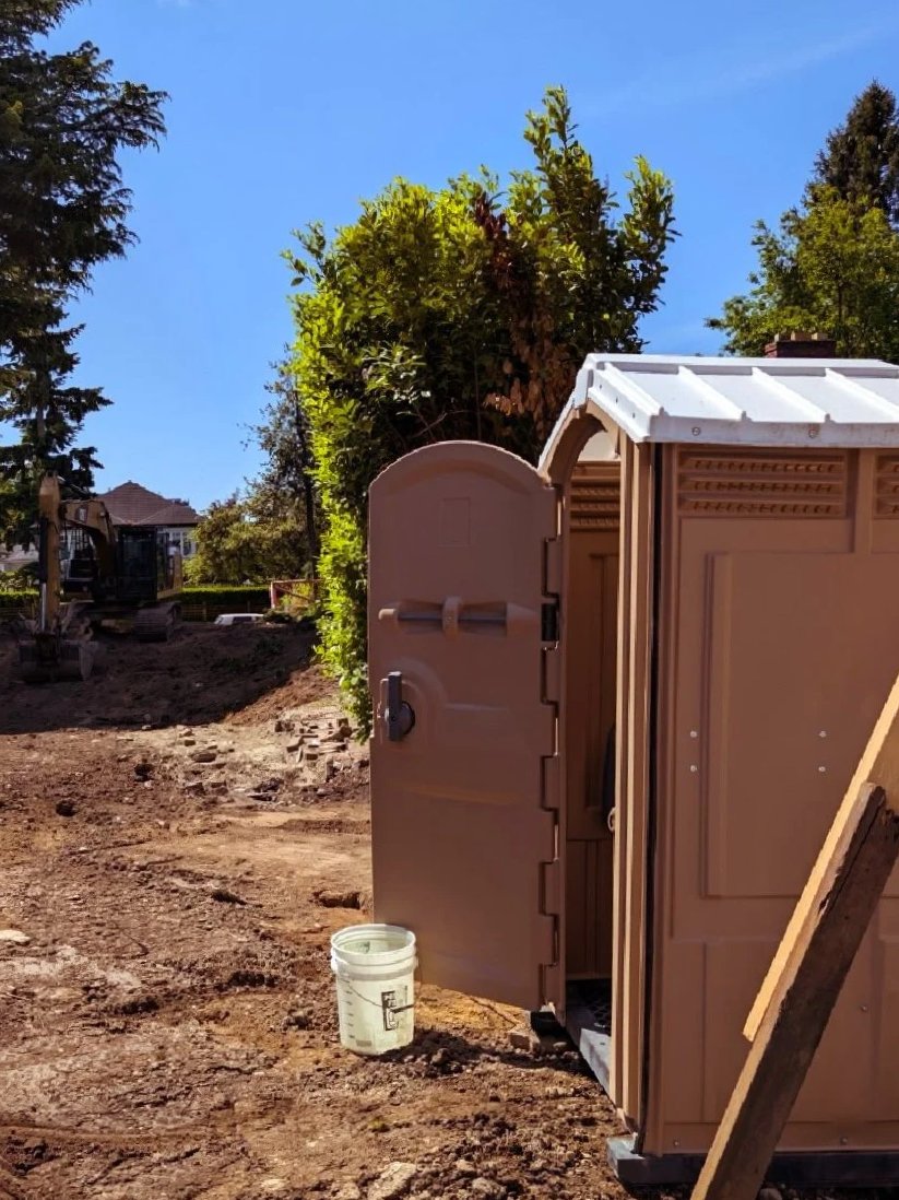 Portable toilet on a construction site with dirt ground, a white bucket in front, and trees and a house in the background.