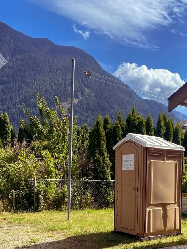 A portable toilet in a grassy yard with a chain-link fence, tall trees, and mountains in the background under a partly cloudy sky.