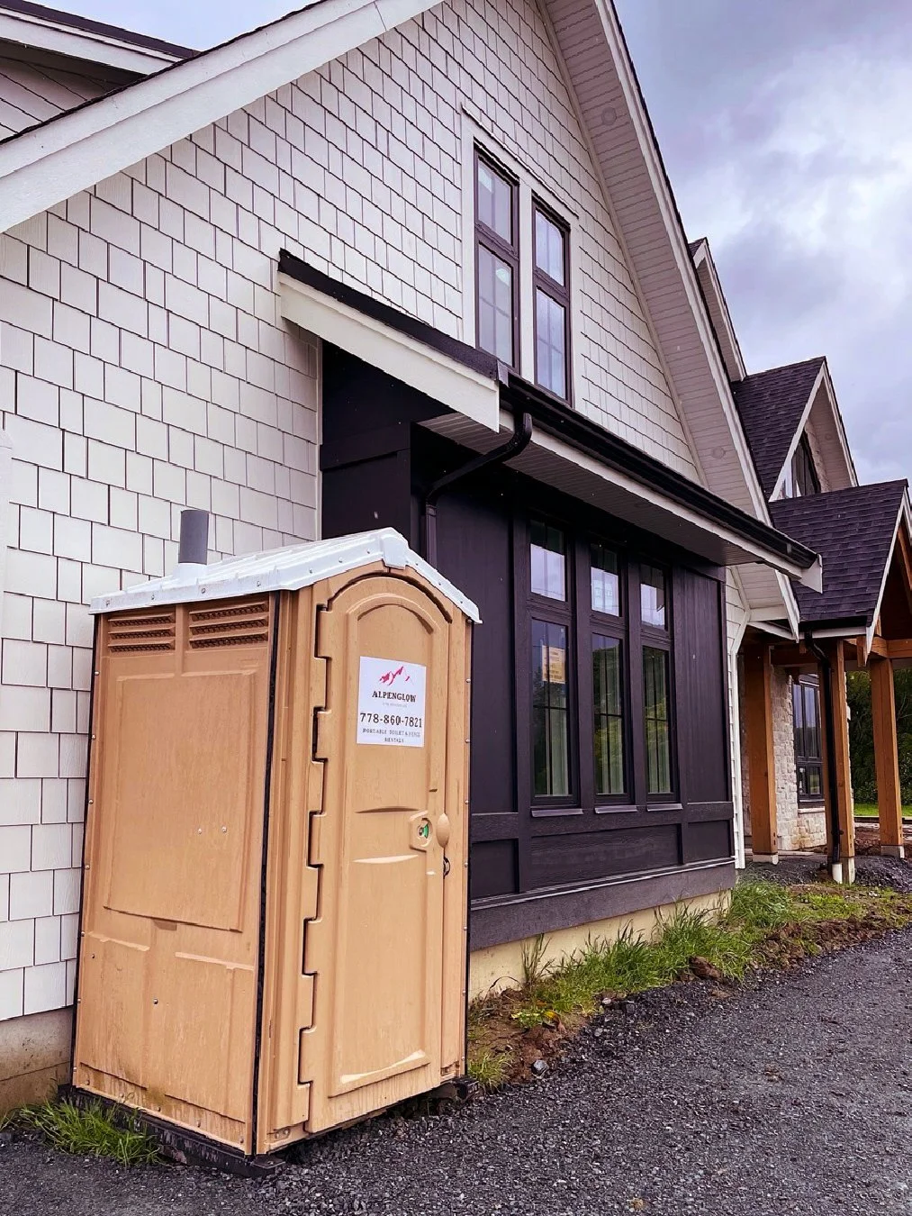 A portable toilet placed outside a house with white shingle siding, black window frames, and a dark roof, on a gravel pathway with some grass nearby.