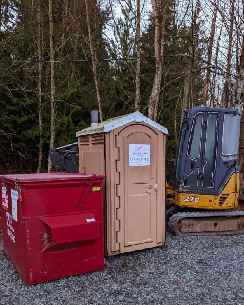 A small portable toilet, a red dumpster, and a small yellow and gray excavator are situated outdoors on a gravel surface with trees and power lines in the background.