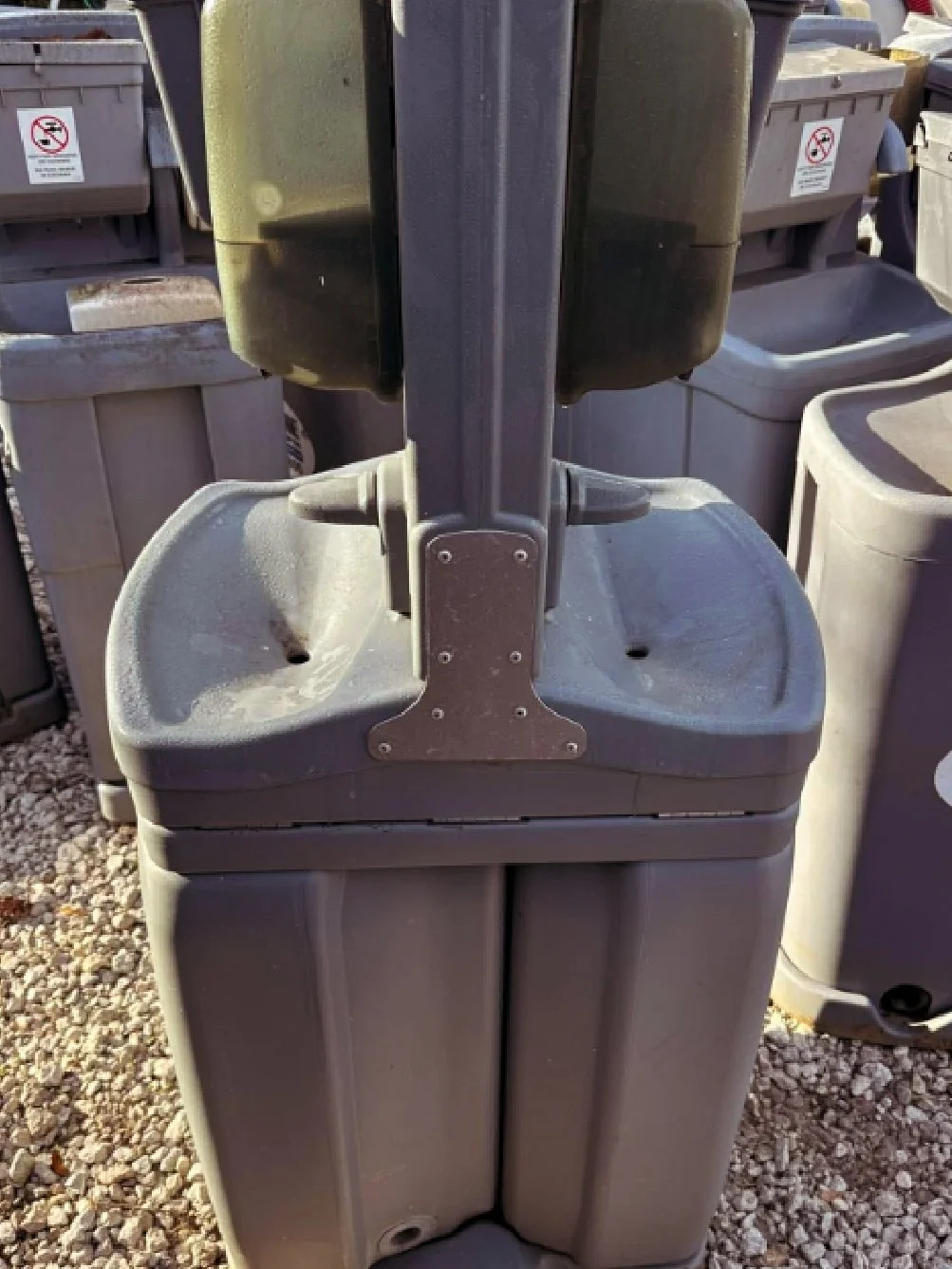 A grey plastic portable hand-washing station on rocky ground under sunlight.