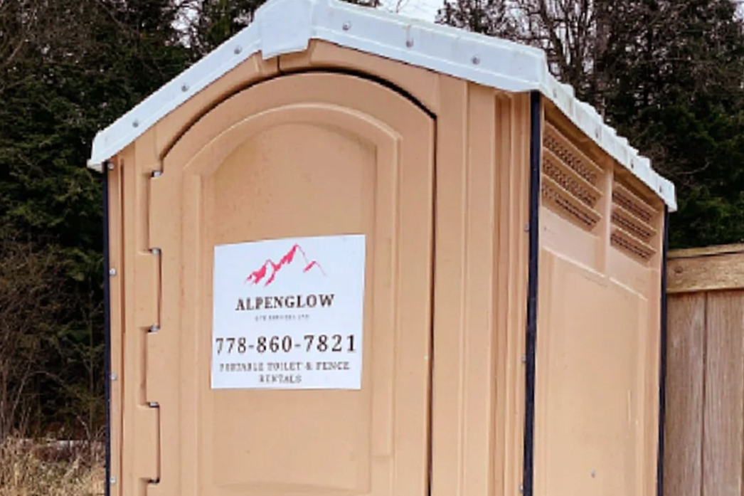 Portable toilet in beige color with a white roof, positioned outdoors near a wooden fence and trees.
