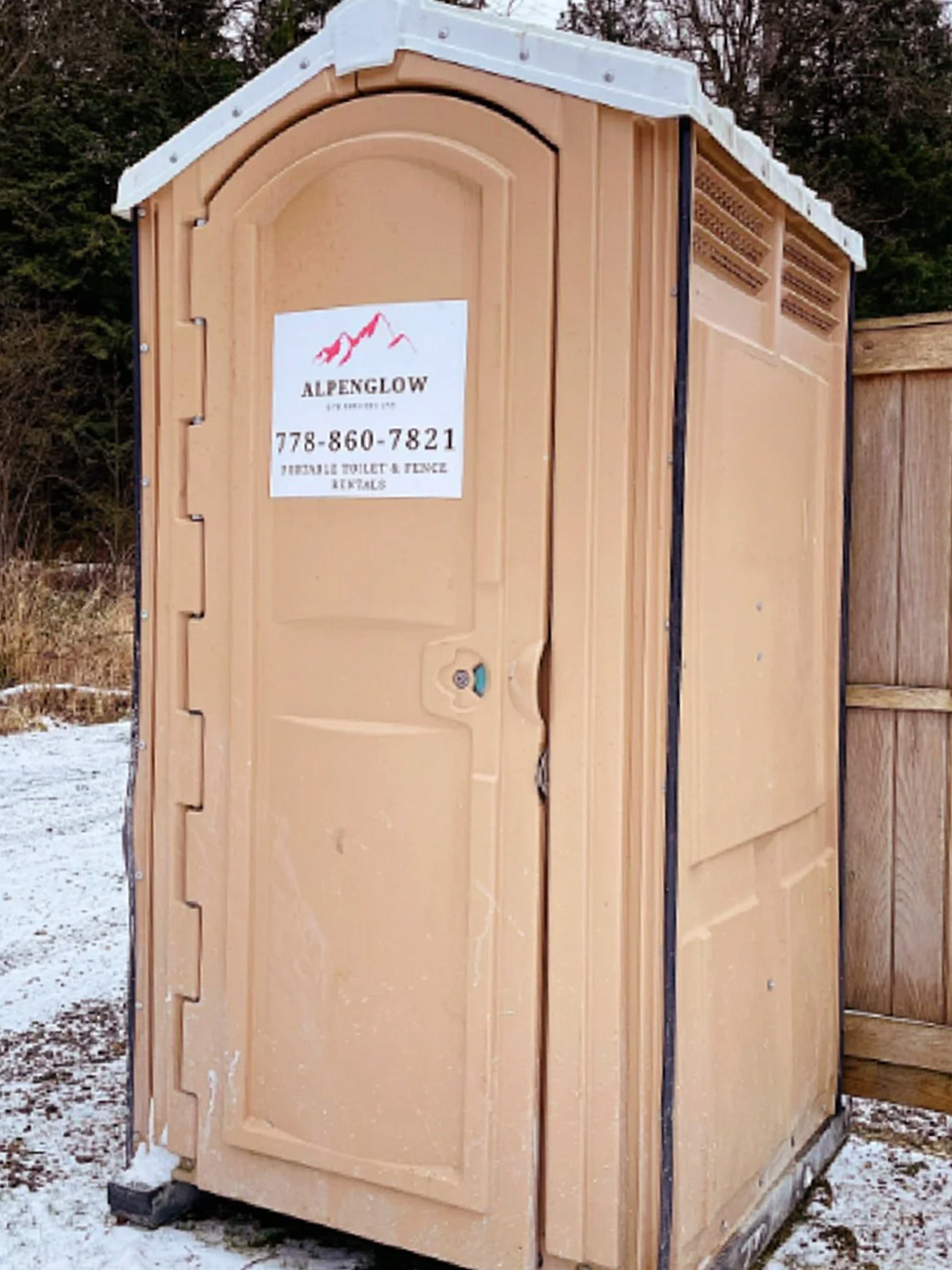 A portable beige toilet with a sign that reads 'Alpenglow, Portable Toilet & Fences Rentals' and a phone number, situated outdoors on snow-covered ground next to a wooden fence, with trees in the background.