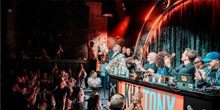 Comedian performs on stage at a comedy club with a panel of other comedians and an audience clapping and cheering.