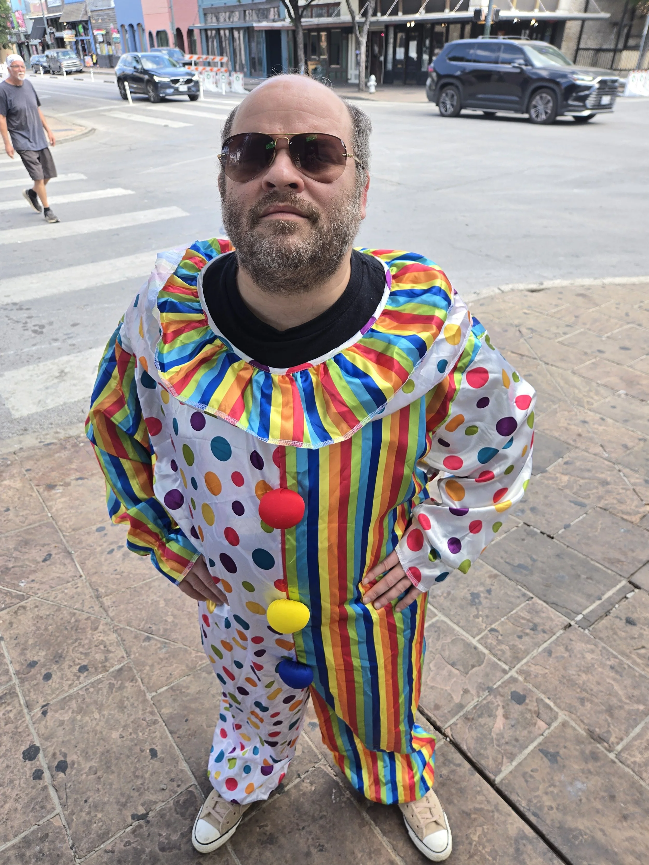 Person dressed as a clown with rainbow-colored polka dot clothing, standing outdoors on a sidewalk in a city, with parked cars and buildings in the background.