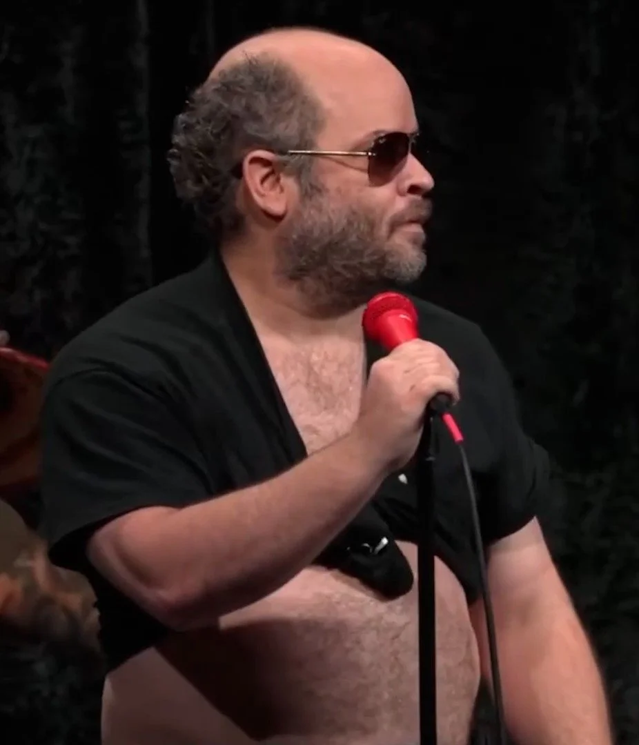 A man with a beard, sunglasses, and a bald head wearing a black shirt, holding a red microphone, with a dark background.